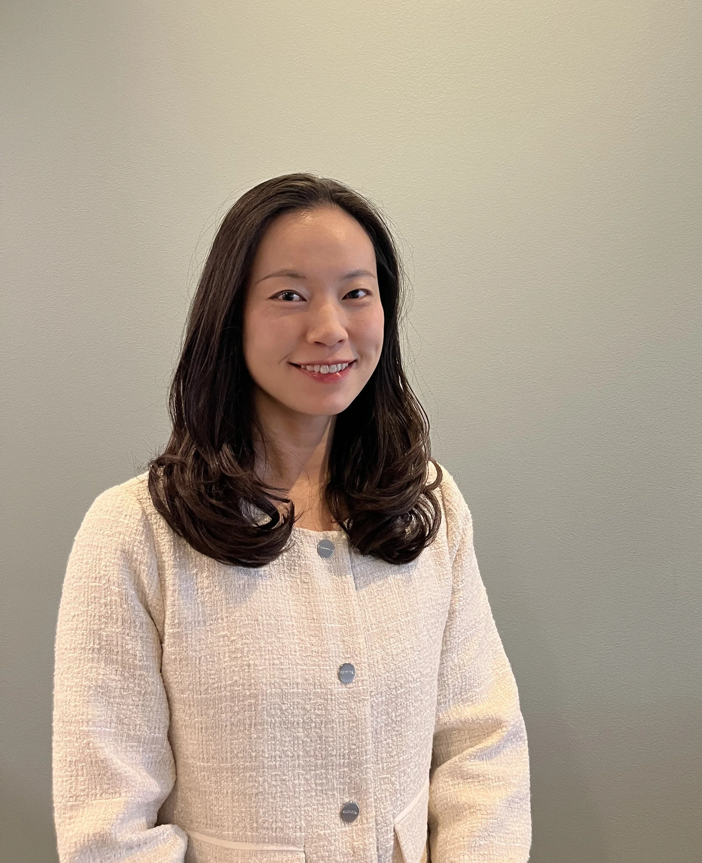A woman with dark brown, wavy hair, smiling, wearing a cream textured jacket with large buttons, standing against a plain beige wall.