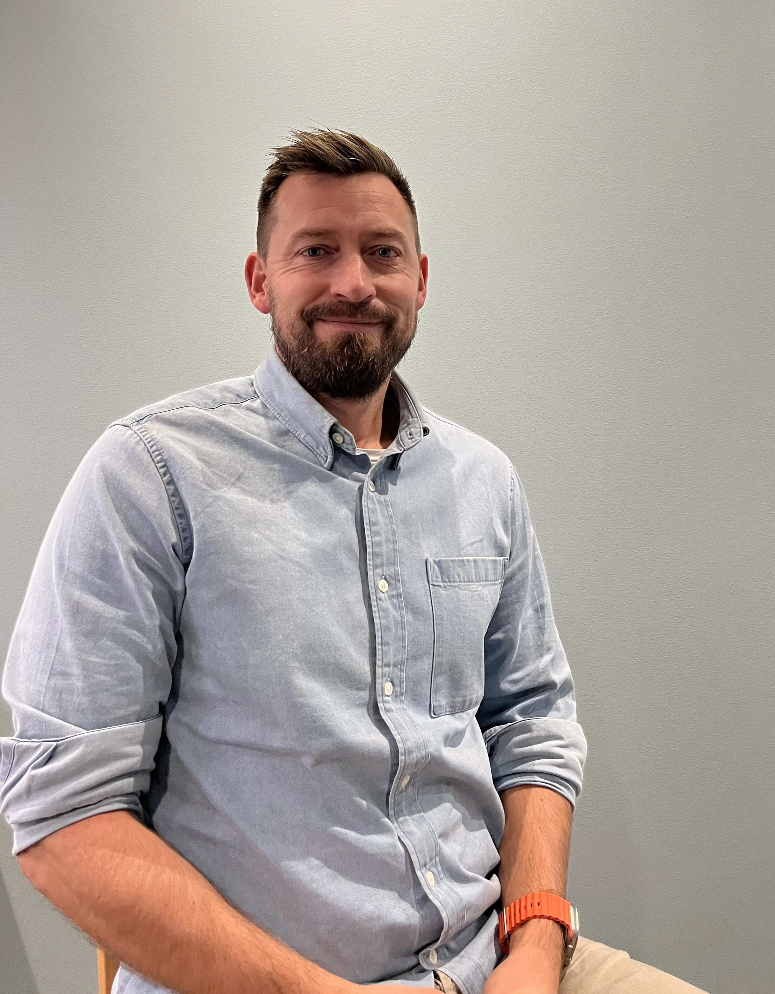 A man with a beard and short dark hair, wearing a light gray button-up shirt and a red watch, sitting against a plain light gray wall.