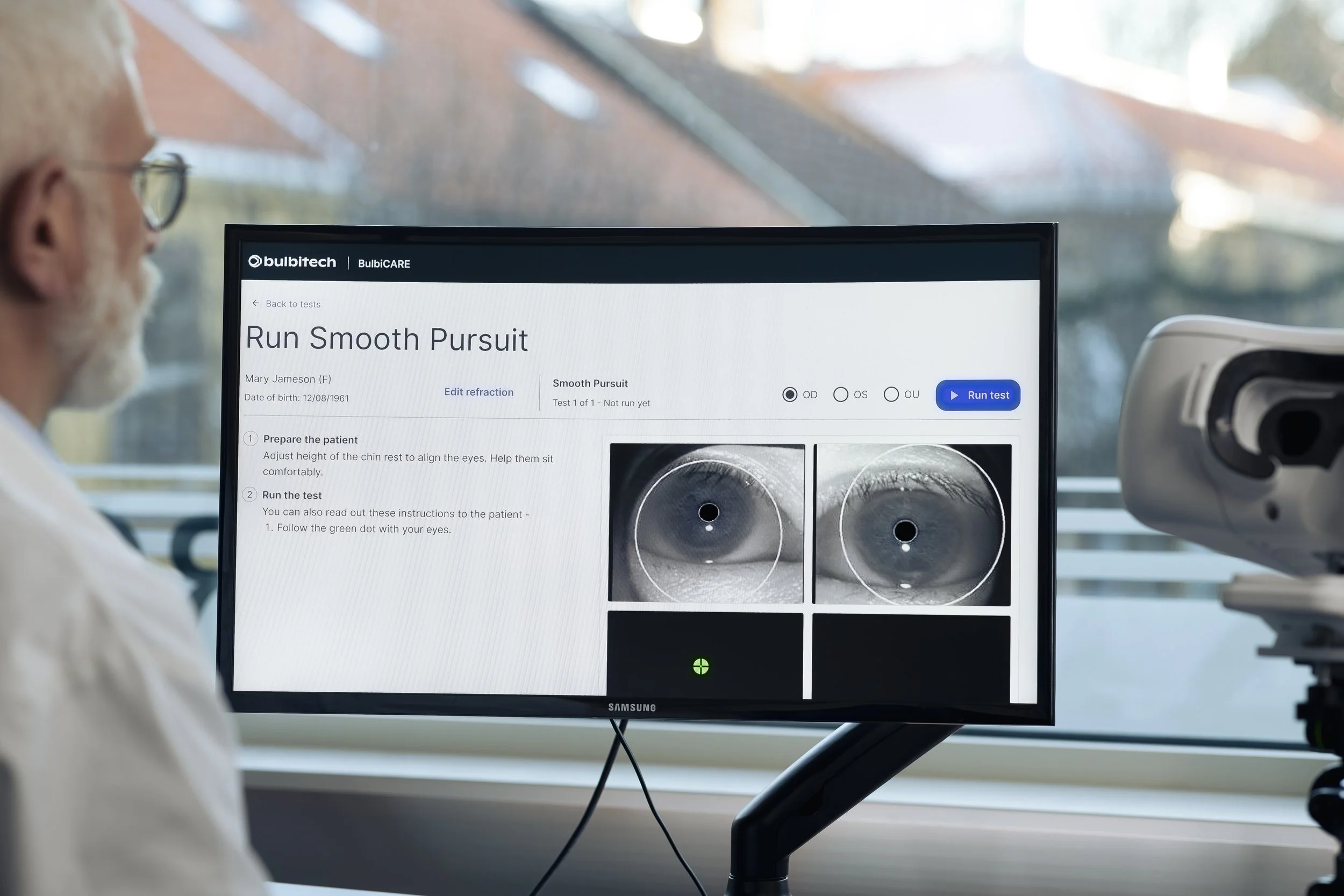 A person in a white lab coat with glasses is looking at a computer monitor displaying an eye test for a procedure called Run Smooth Pursuit. The screen shows instructions and two close-up black-and-white images of eyes with targets to follow for testing.