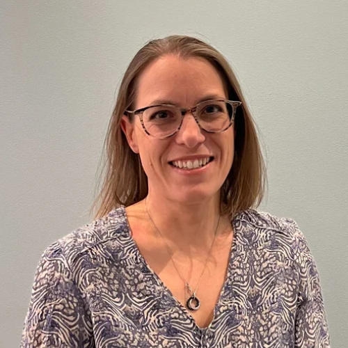 A woman with glasses smiles at the camera against a plain wall.