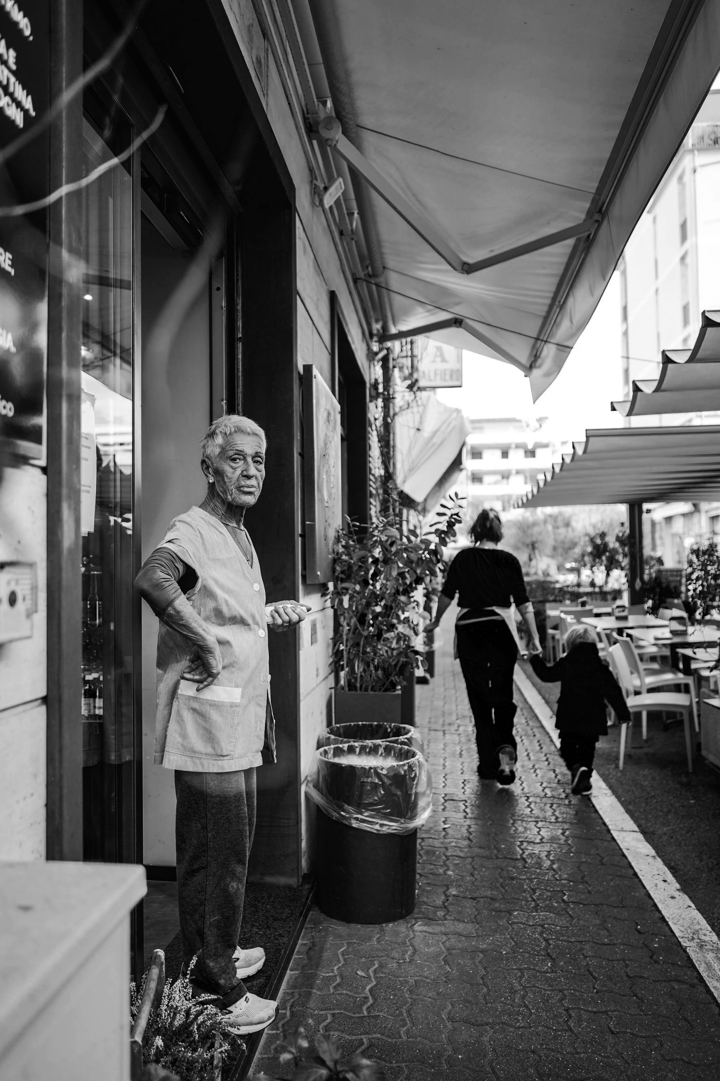 Una donna anziana con i capelli bianchi e vestiti comodi si trova davanti a un negozio o ristorante, mentre sullo sfondo una donna con un bambino si allontanano lungo un marciapiede all'aperto con sedie e ombrelloni. Pizzeria Alfiero Orto di Alfiero