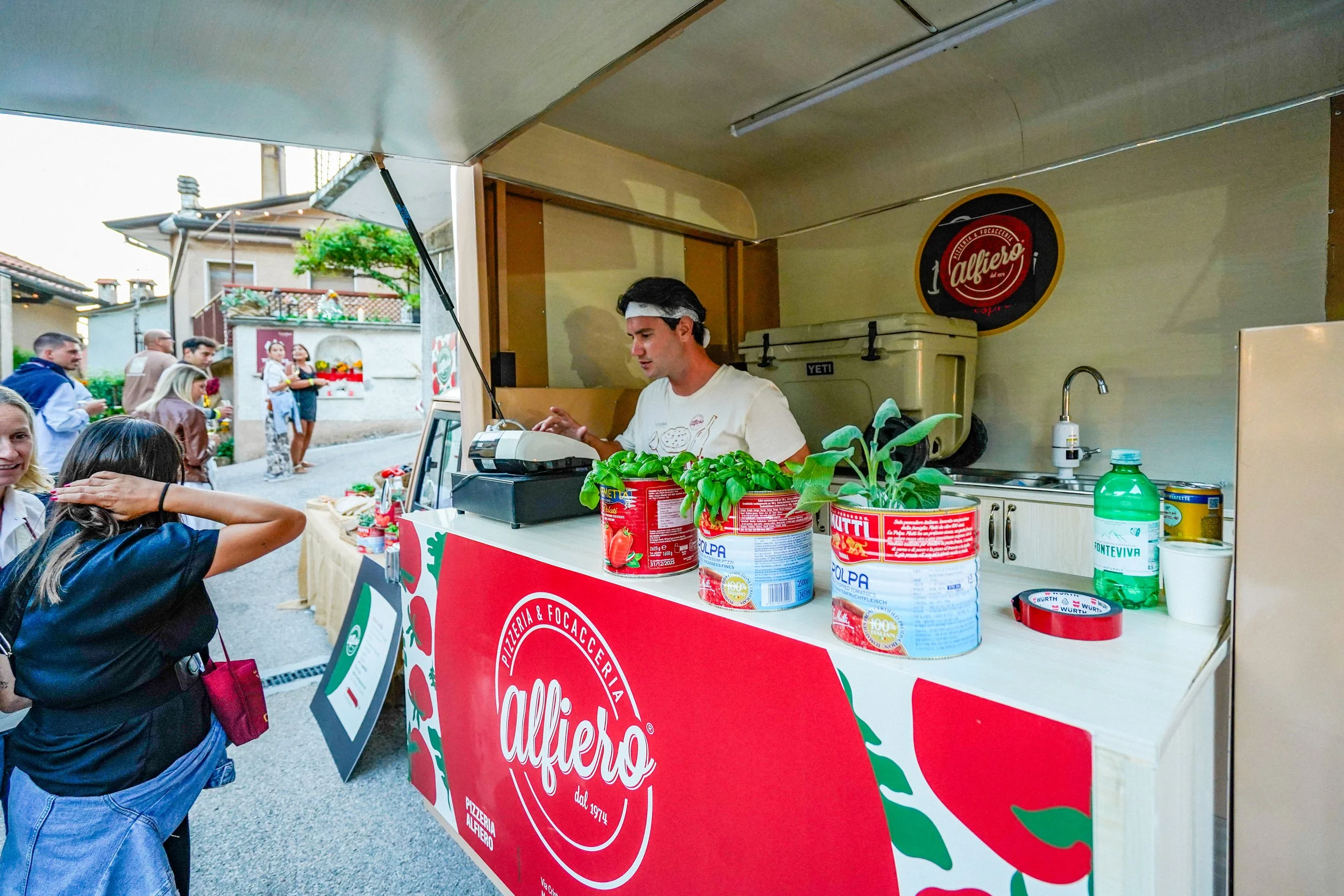 Un giovane venditore alla cassa di un chiosco di pizza con il logo 'Alfio' serve clienti in un evento all'aperto. Pizzeria Alfiero Orto di Alfiero Marina di Massa.