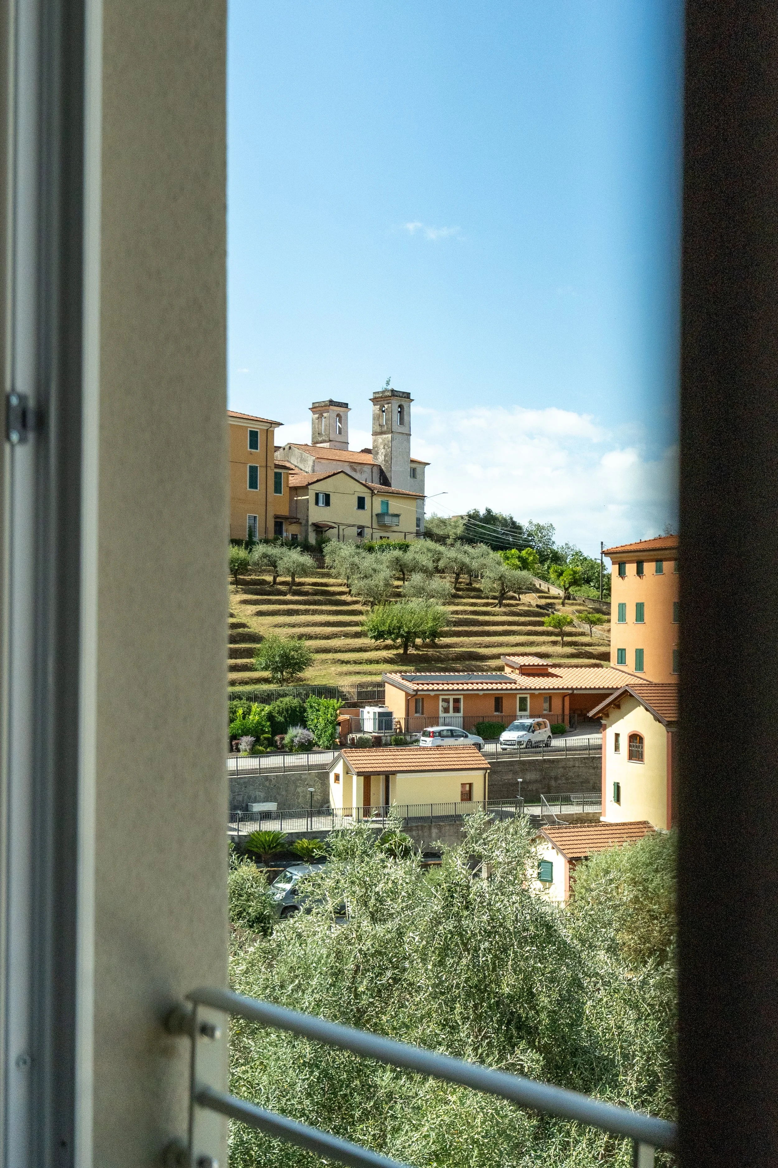 Vista su un paesaggio collinare con case colorate e un serie di torri di un edificio storico sullo sfondo, visibile attraverso una finestra. Residenze Volpigliano Massa