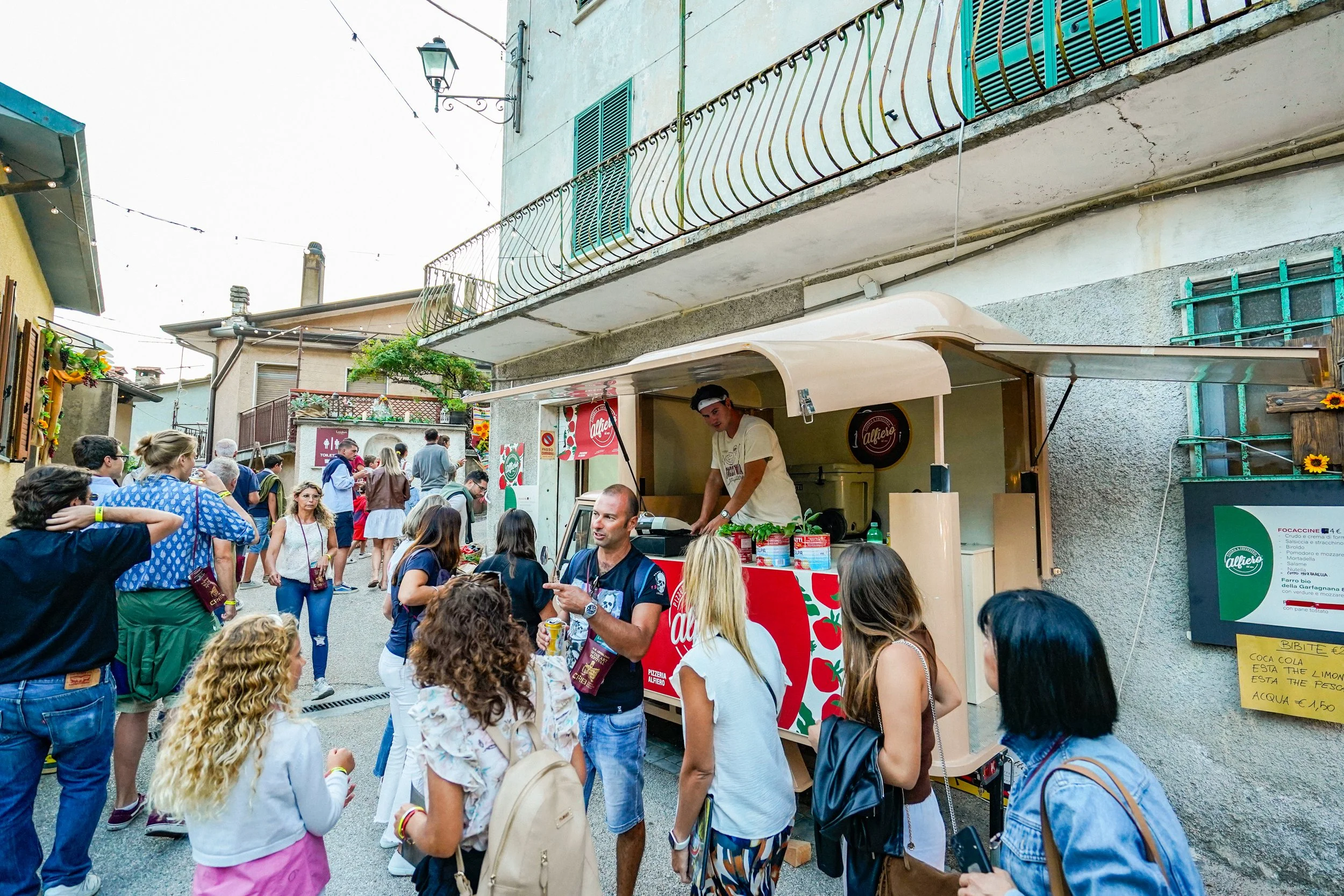 Stands di cibo di strada con molte persone che fanno fila, una piccola cucina mobile con un cuoco che prepara cibo, in una strada affollata con edifici colorati. Pizzeria Alfiero Orto di Alfiero Marina di Massa.