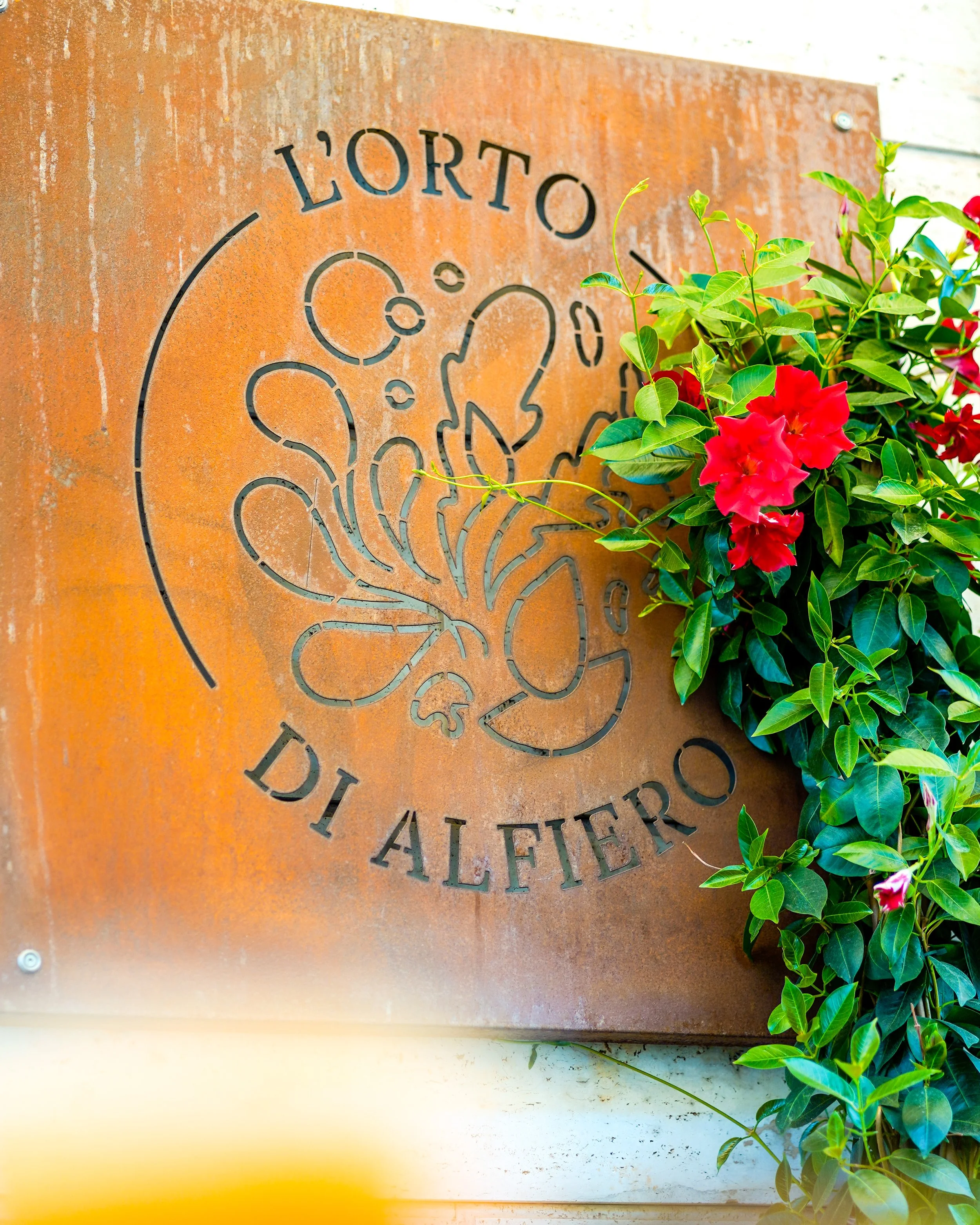 Pannello in metallo con scritta 'L'Orto di Alfiero' e un disegno stilizzato di un fiore, circondato da foglie verdi e fiori rossi. Pizzeria Alfiero Orto di Alfiero Marina di Massa.