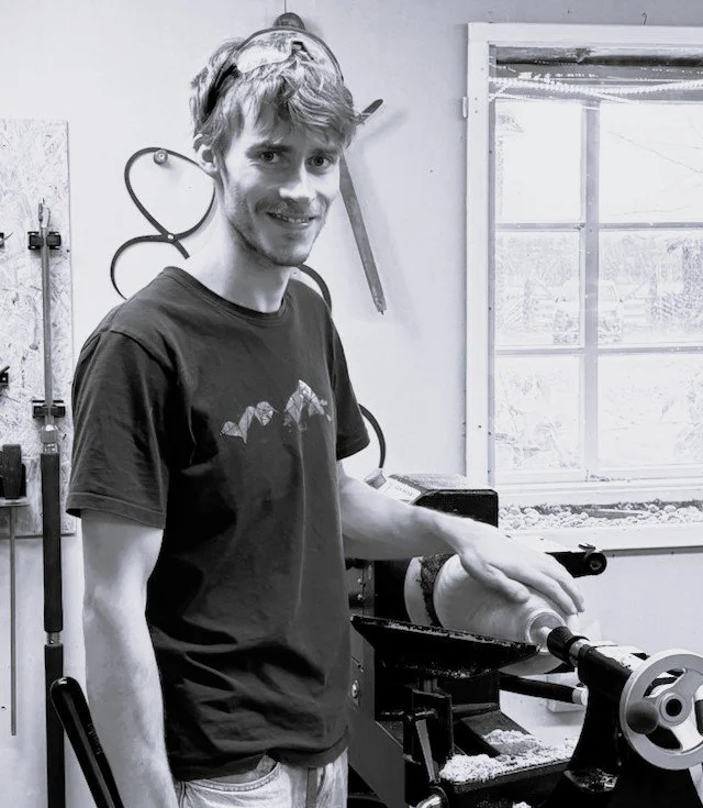 En ung mann i svart t-skjorte som bruker en trebearbeidingsmaskin, i et verksted med verktøy og et vindu i bakgrunnen.