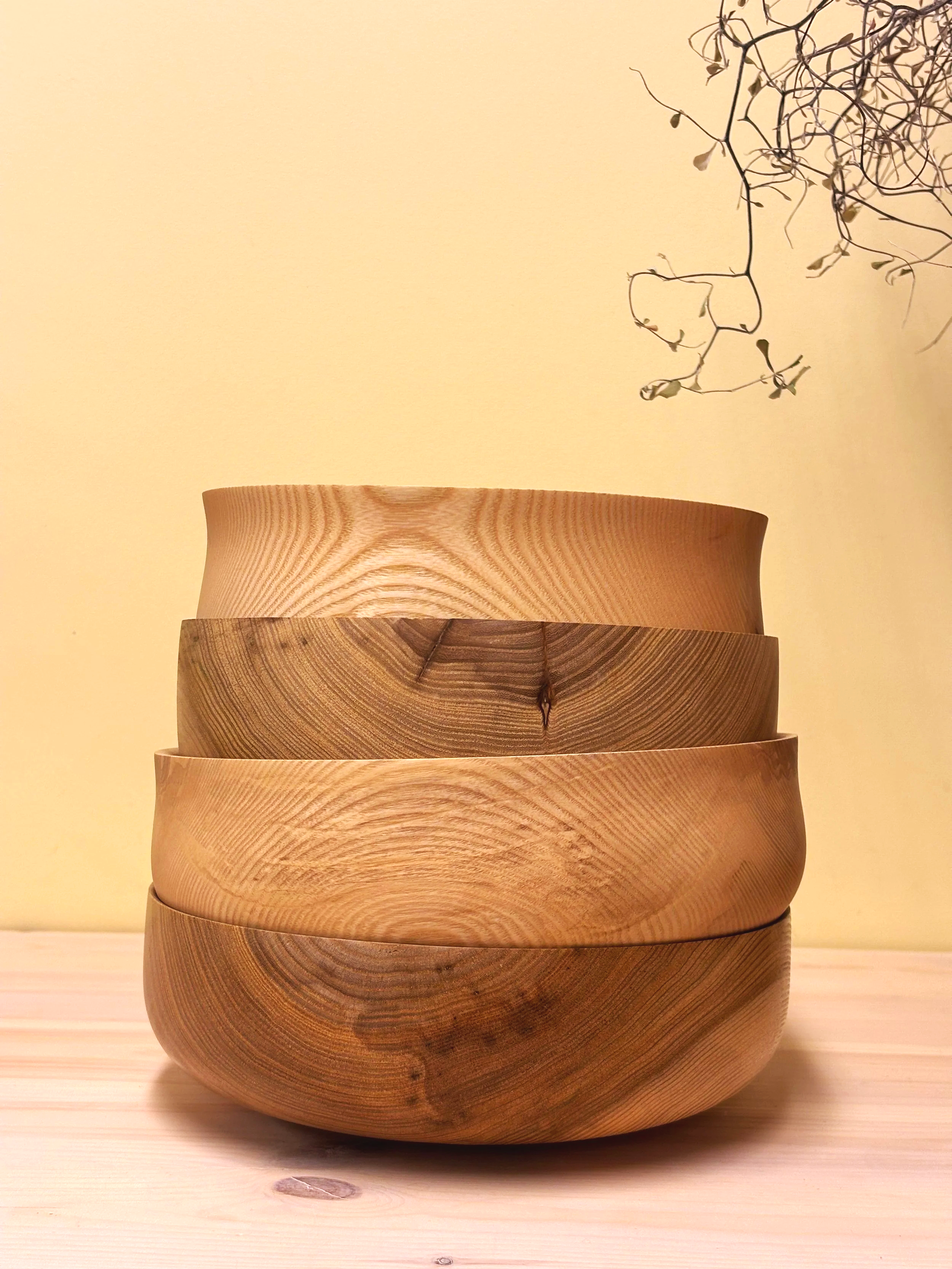 Fire trerlignende skåler stablet oppå hverandre på et lyst trebord, med en svak beige vegg i bakgrunnen og en grein med små, tørre blader i øvre høyre hjørne.