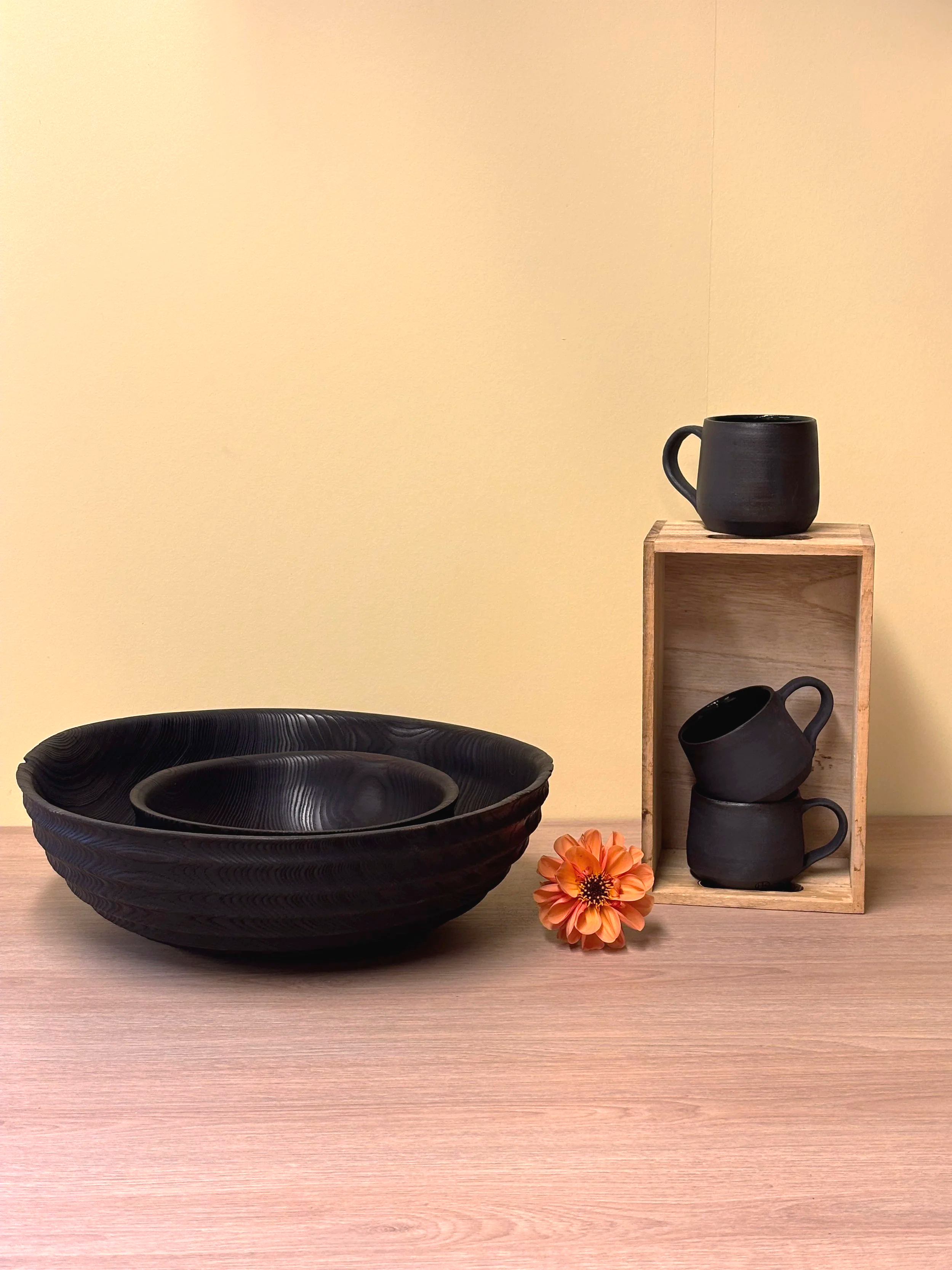 En samling svarte keramiske kopper og skåler plassert på en lys trebord med en lys gul bakgrunn. Det er også en oransje blomst foran koppene.