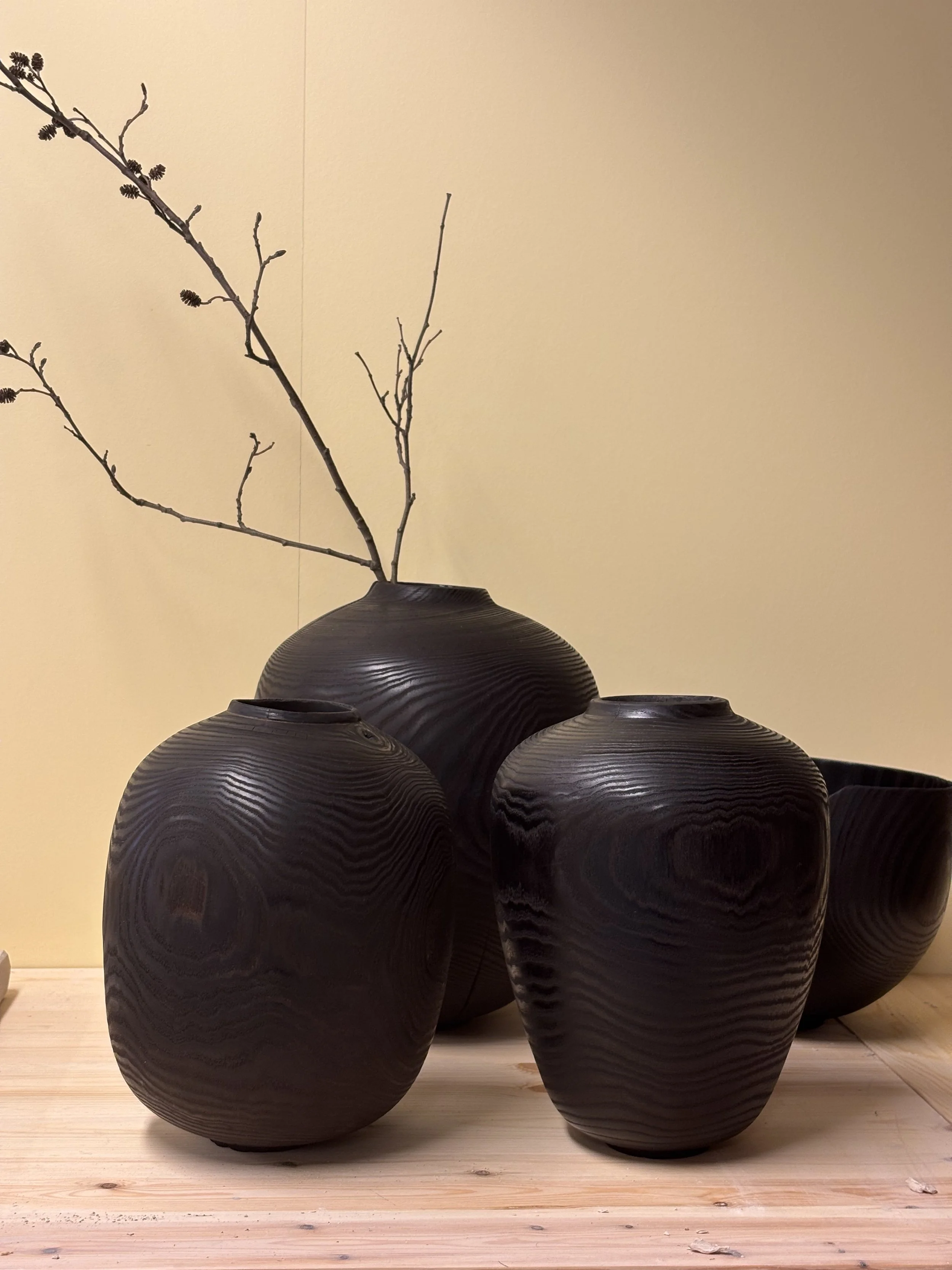 Fire svarte keramikkvaser med tre grener uten blader plassert på en lys trebord, mot en beige vegg.