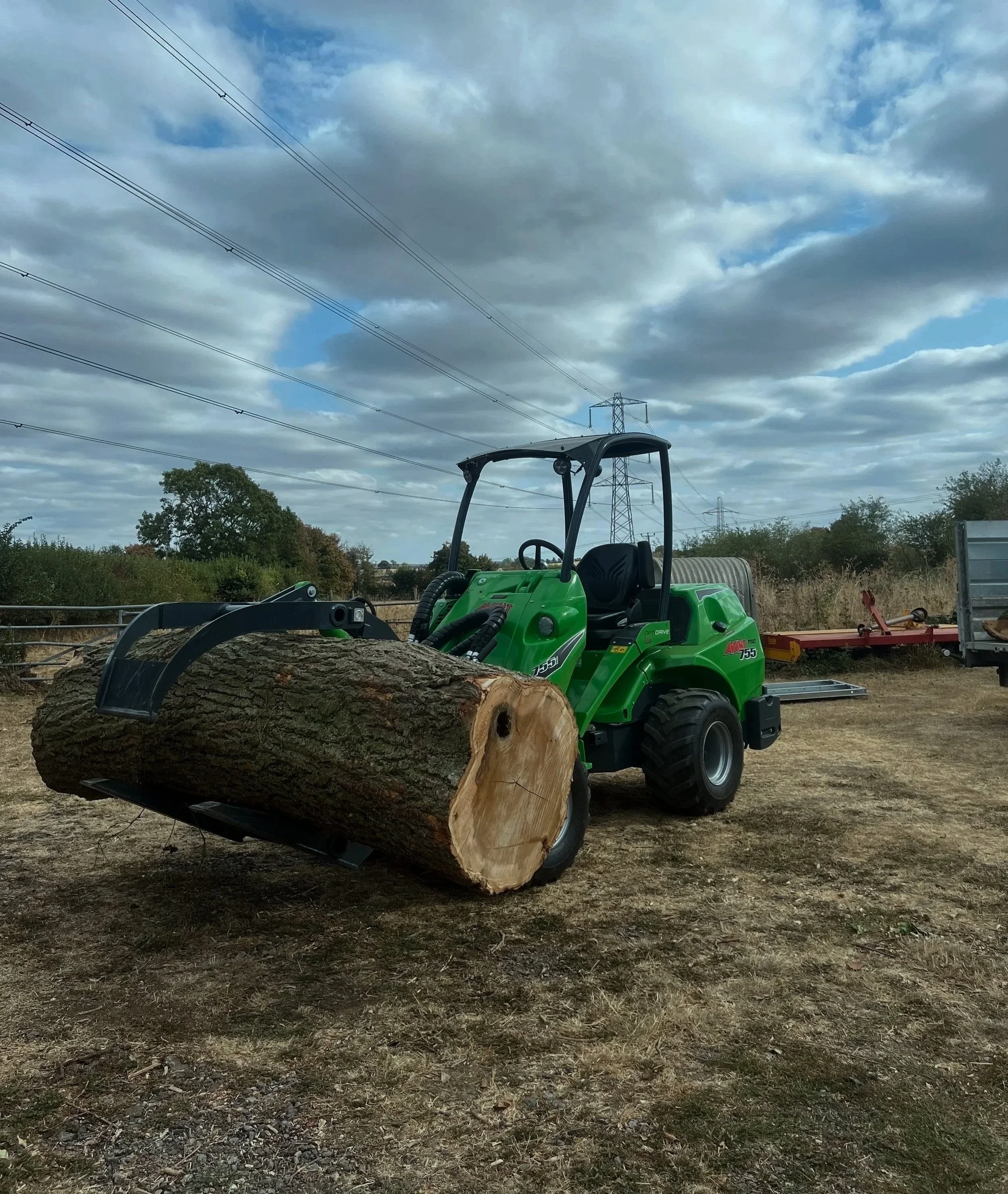 A green compact loader with a large log in the bucket, set outdoors on a dirt patch with trees, power lines, and cloudy sky in the background.