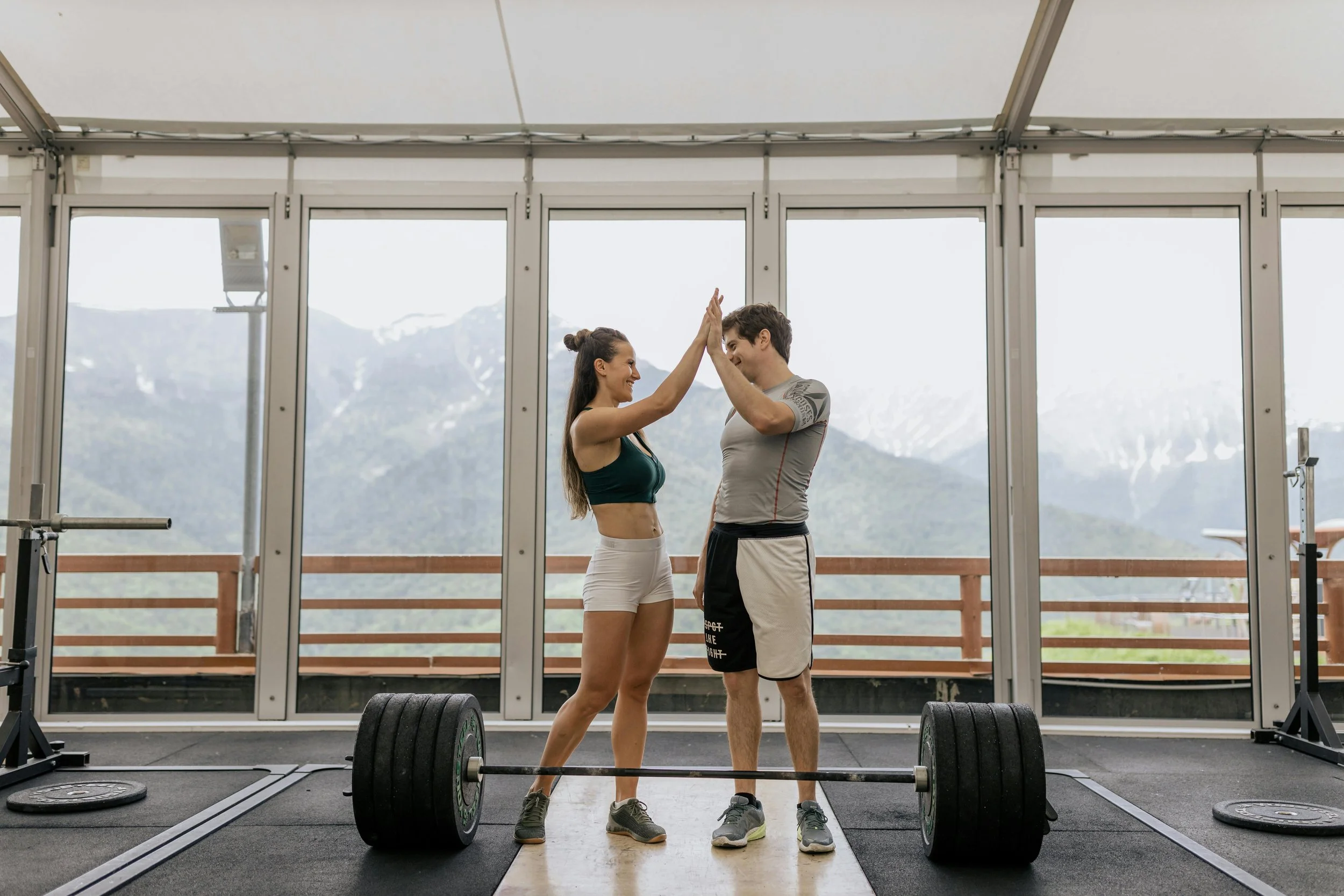 Pareja en gimnasio dándose un toque de manos en señal de celebración, con pesas en el suelo y fondo de montañas.