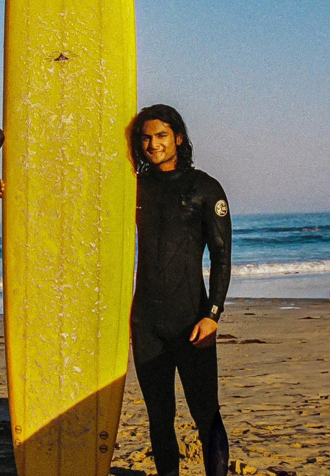 Thisal standing on the beach next to a yellow surfboard, smiling at the camera, with the ocean in the background.
