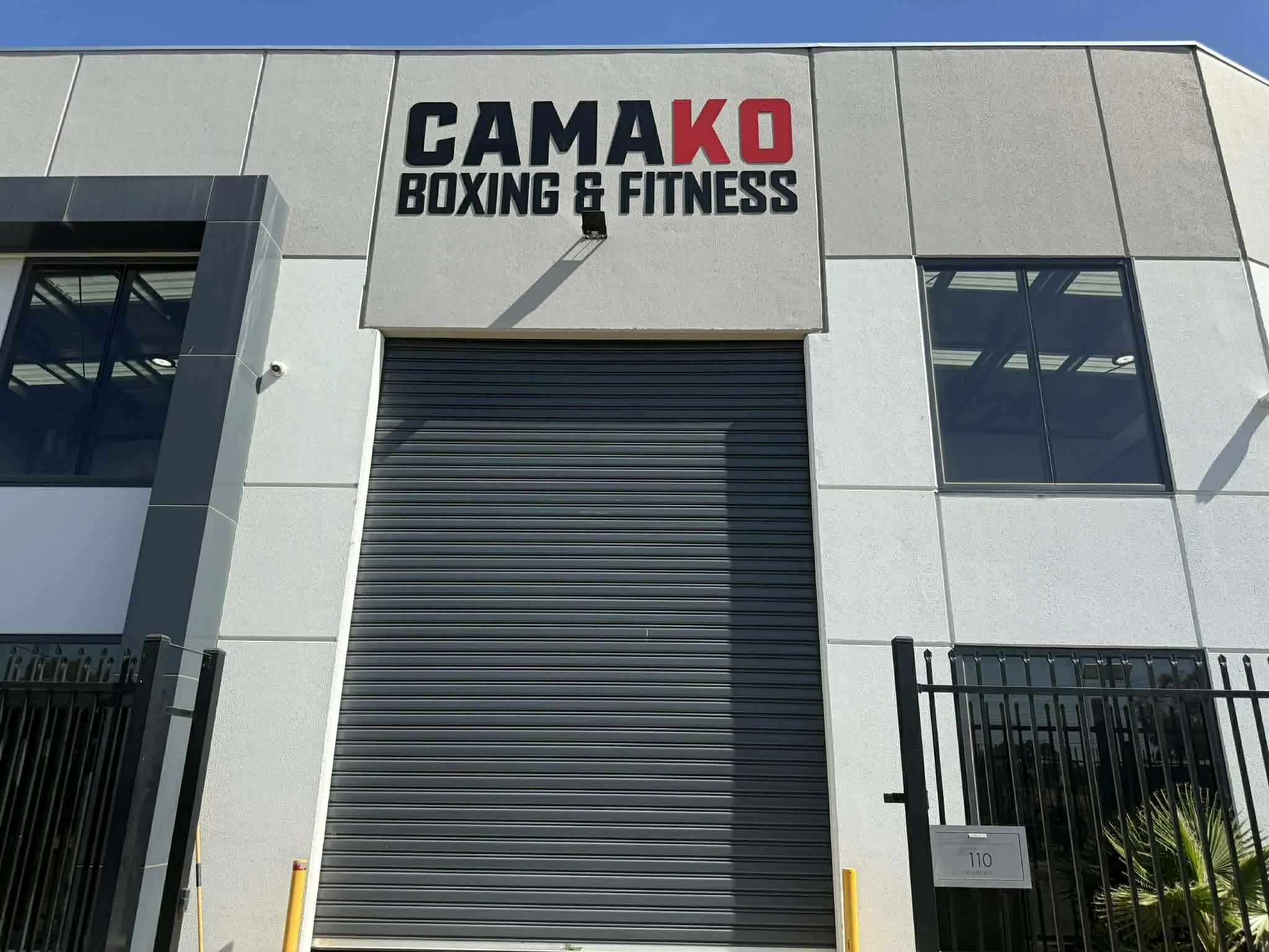 Front exterior of a gym with a sign that reads "CAMA KO Boxing & Fitness" and a large closed rolling door, fenced area, and windows.