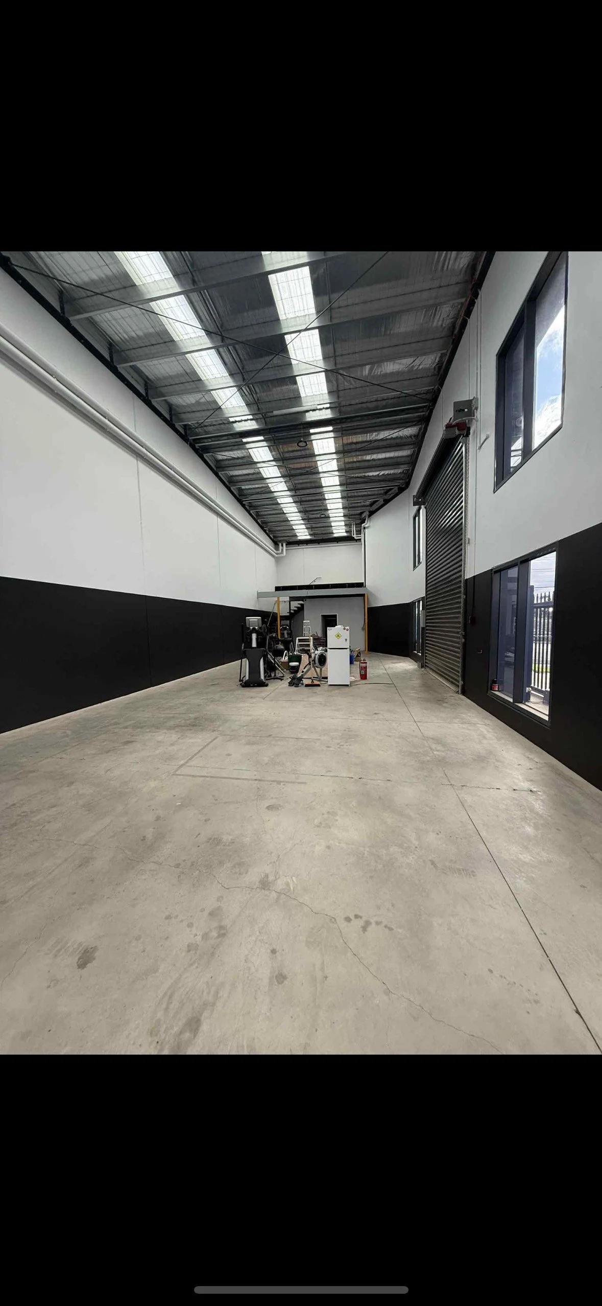 An empty industrial warehouse with a polished concrete floor, white walls with black lower half, large windows, and a high ceiling with exposed metal roofing. There are various machinery and equipment near the back wall, including a fire extinguisher and a roll-up door.