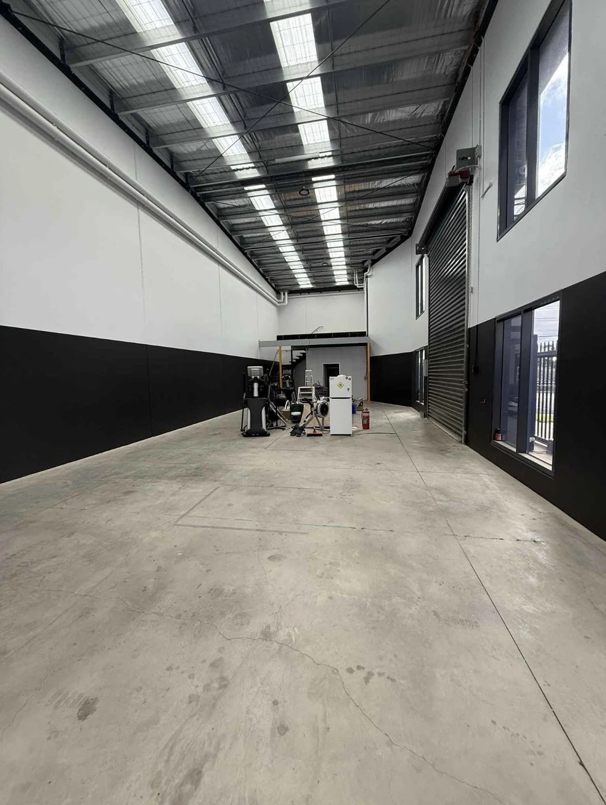 An empty industrial or warehouse space with a concrete floor, high ceiling with translucent panels, black and white walls, and some equipment and fire extinguisher at the back.