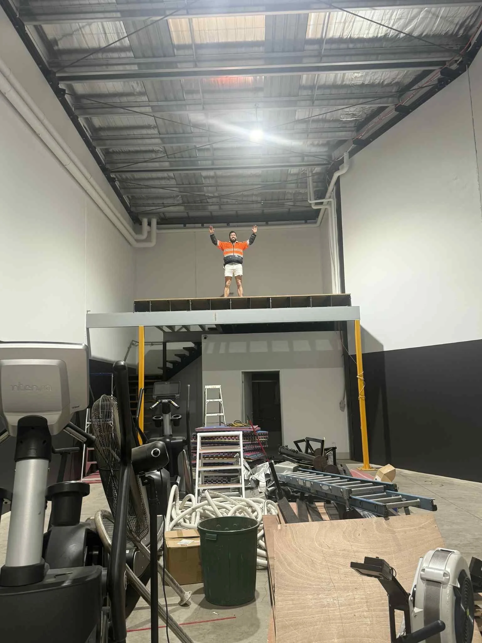 A person standing on a raised platform with arms raised inside a large industrial space with high ceilings, metal roof, and construction equipment and materials scattered on the ground.