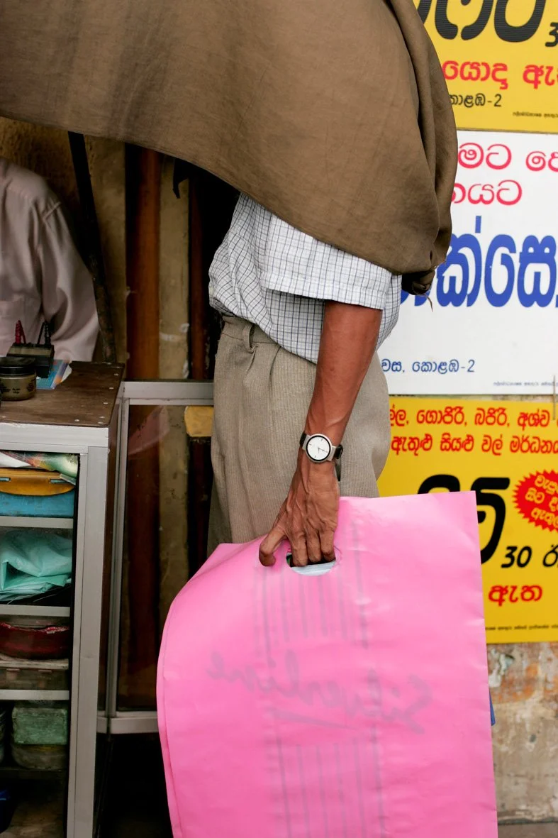 Homme Sri Lankais dans une rue du Sri Lanka, tenant dans sa main un sac en plastique rose vif. Sa tête est cachée sous une tenture. 