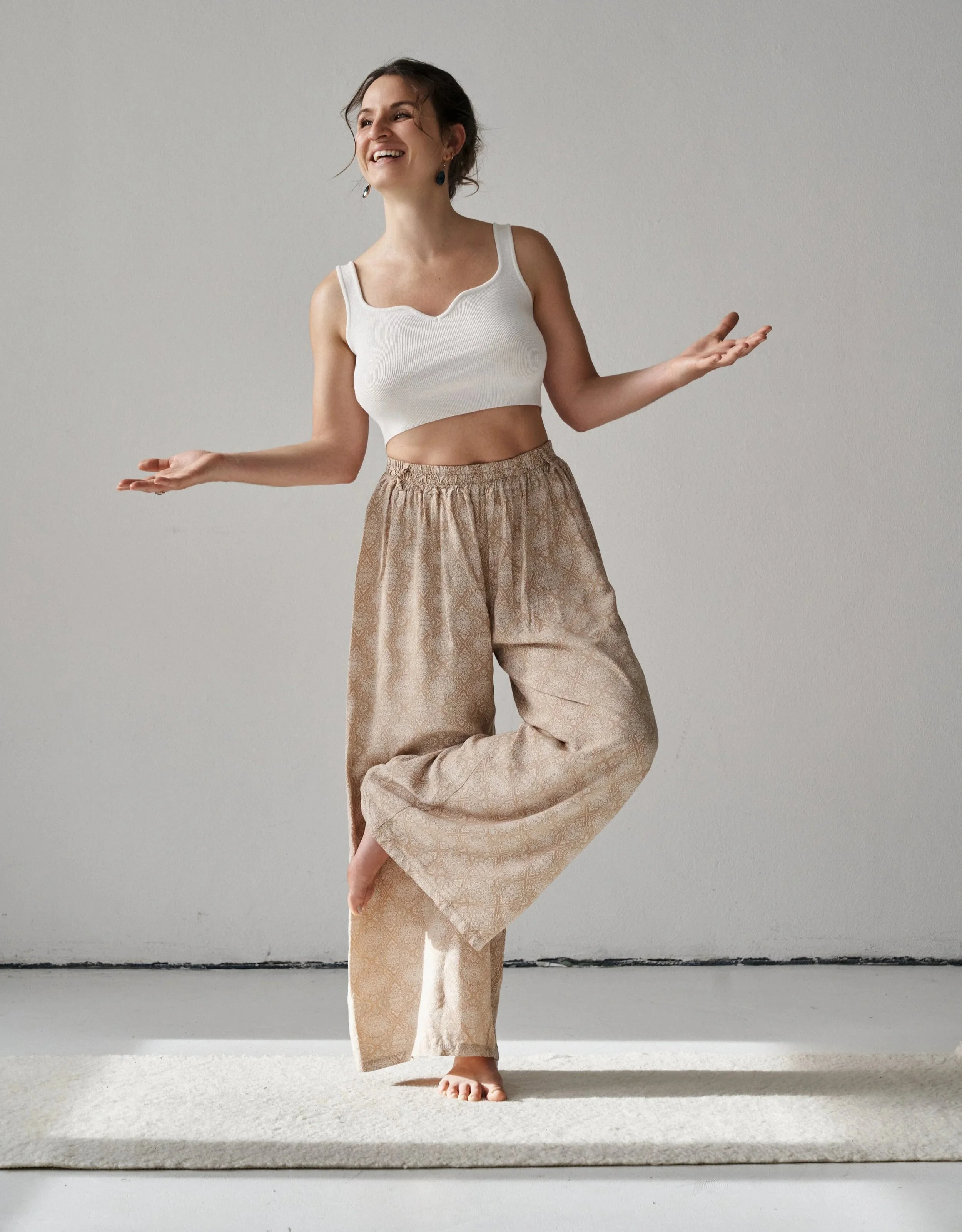 Frau steht auf einem Teppich und übt Yoga, lächelnd mit ausgebreiteten Armen und einem Bein auf der anderen Oberschenkel, trägt weiße Top und weite beigen Hosen.