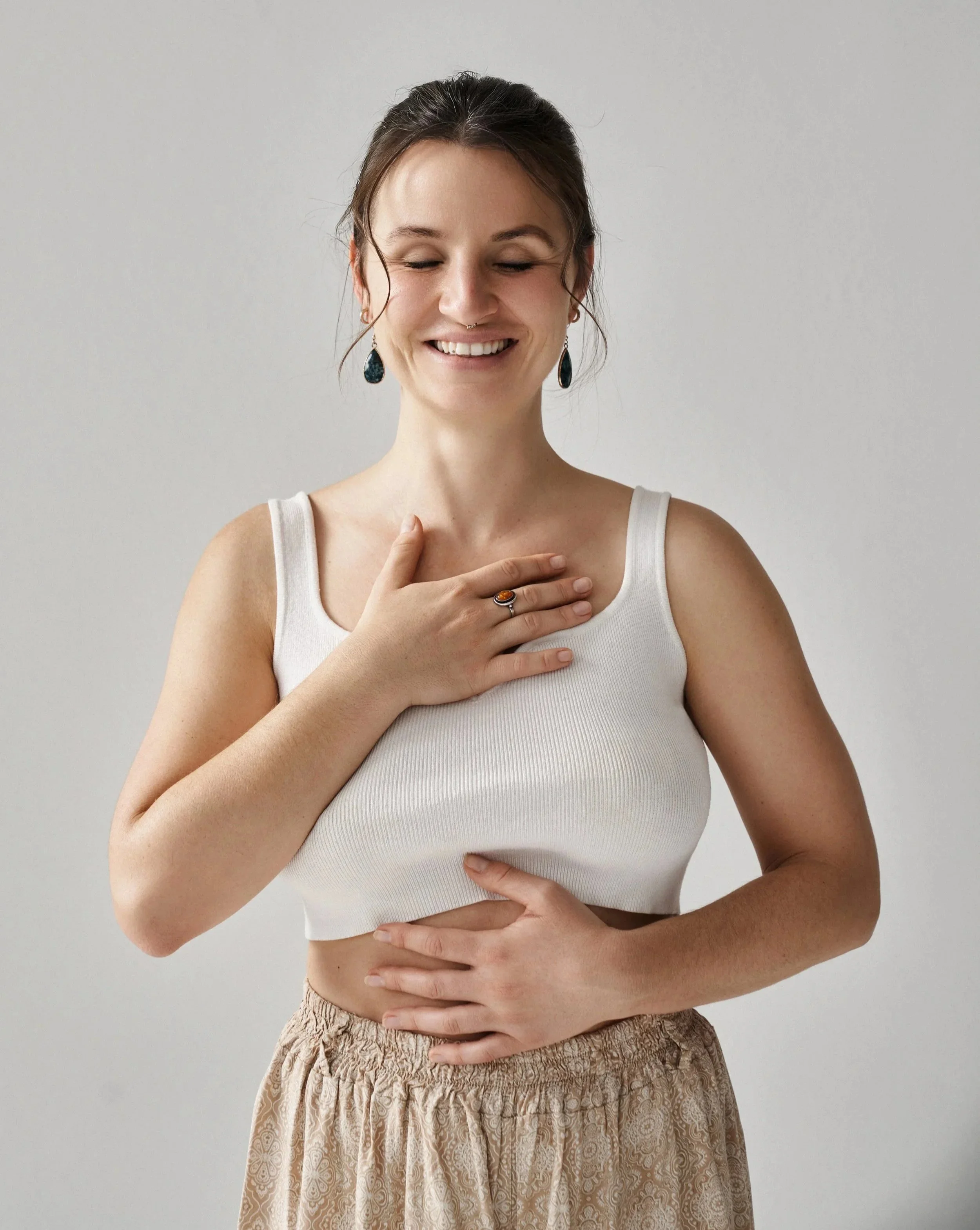 Franziska Riedl, Yoga Lehrerin, lächelt mit geschlossenen Augen in ihr offenes Herz, trägt ein weißes Oberteil und bequeme Hose, eine Hand auf der Brust, die andere auf dem Bauch, die dreiteile Atmung oder yogische Atmung, vor weißem Hintergrund