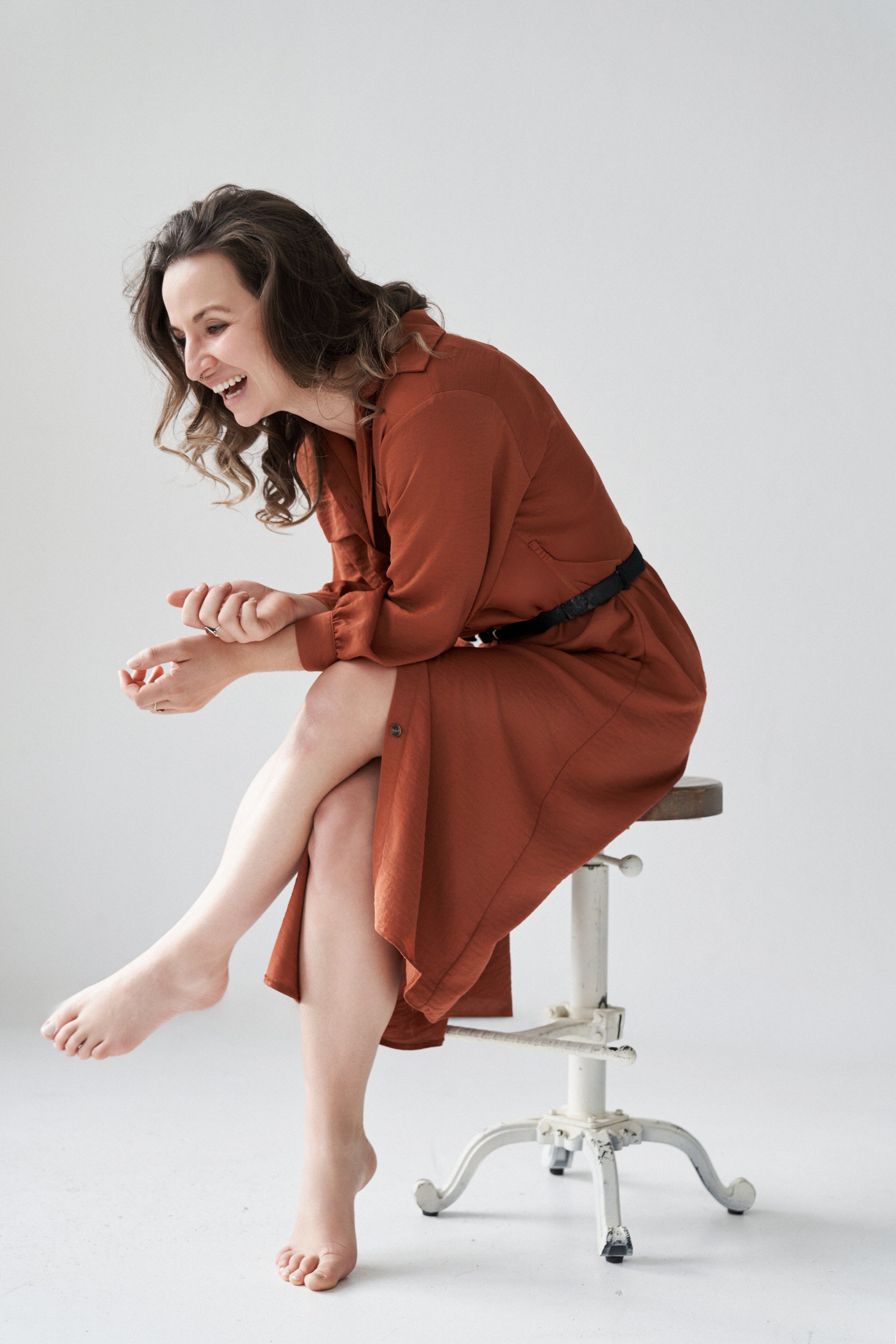 Franziska Riedl in braun rotem Kleid sitzt auf einem Hocker, glücklich lachend in ihr Herz hinein und hat ihre Beine überkreuzt, sie ist barfuß und trägt offene Haare mit Locken für Leichtigkeit, Selbstbewusstsein und Lebensfreude vor weißer Wand