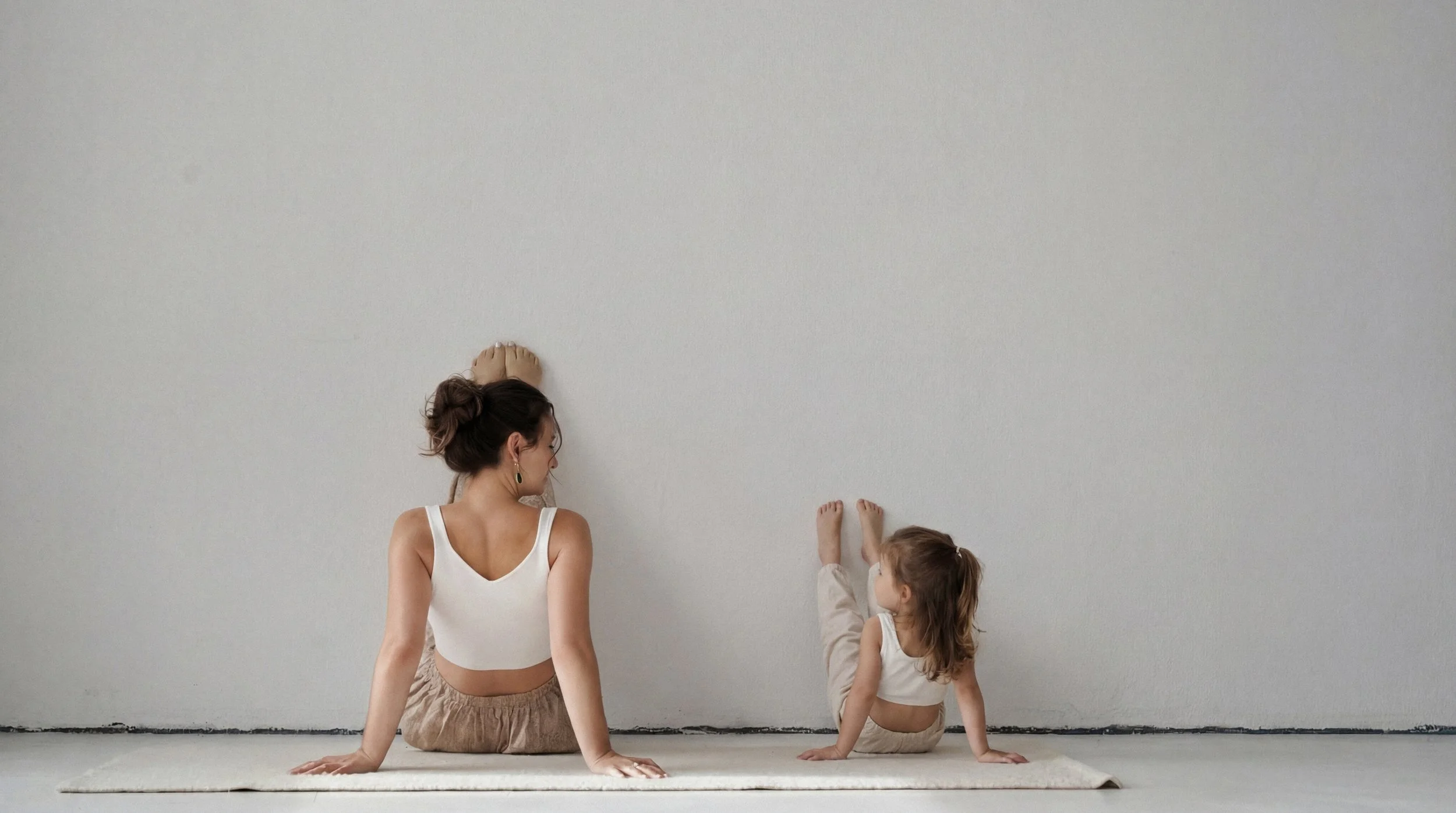 Eine Frau und ein kleines Mädchen, beide in weißen sportlichen Outfits, sitzen auf einem Teppich vor einer weißen Wand, üben Yoga oder Dehnübungen gemeinsam.