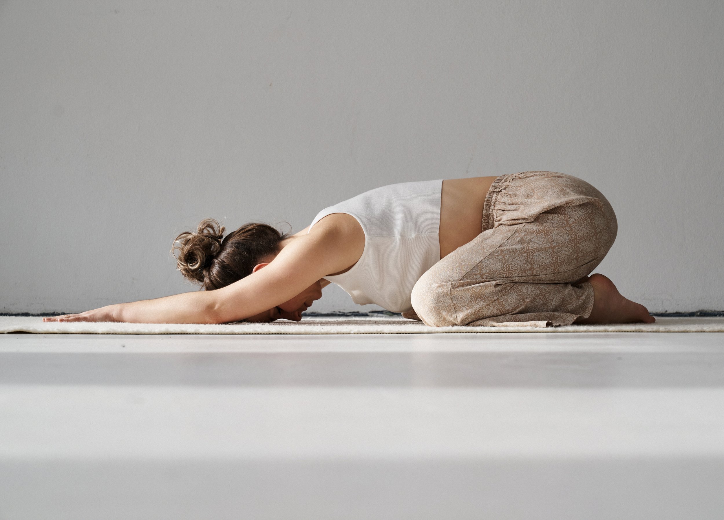 Frau macht Yoga in der Kindhaltung auf einem weißen Teppich vor einer weißen Wand.