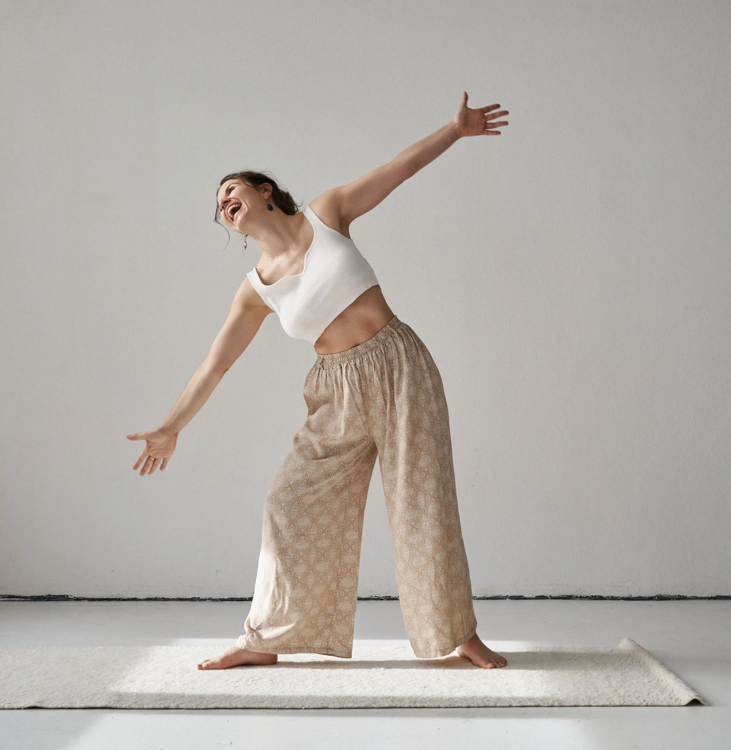 Franziska Riedl in lässiger Kleidung beim Tanzen und der Kriegerhaltung Asana auf einem weißen Teppich in einem leeren Raum für Leichtigkeit, Spaß und Lebensfreude mit offenen Armen und offenem Herzen, bereit persönlichem Wachstum und Transformation