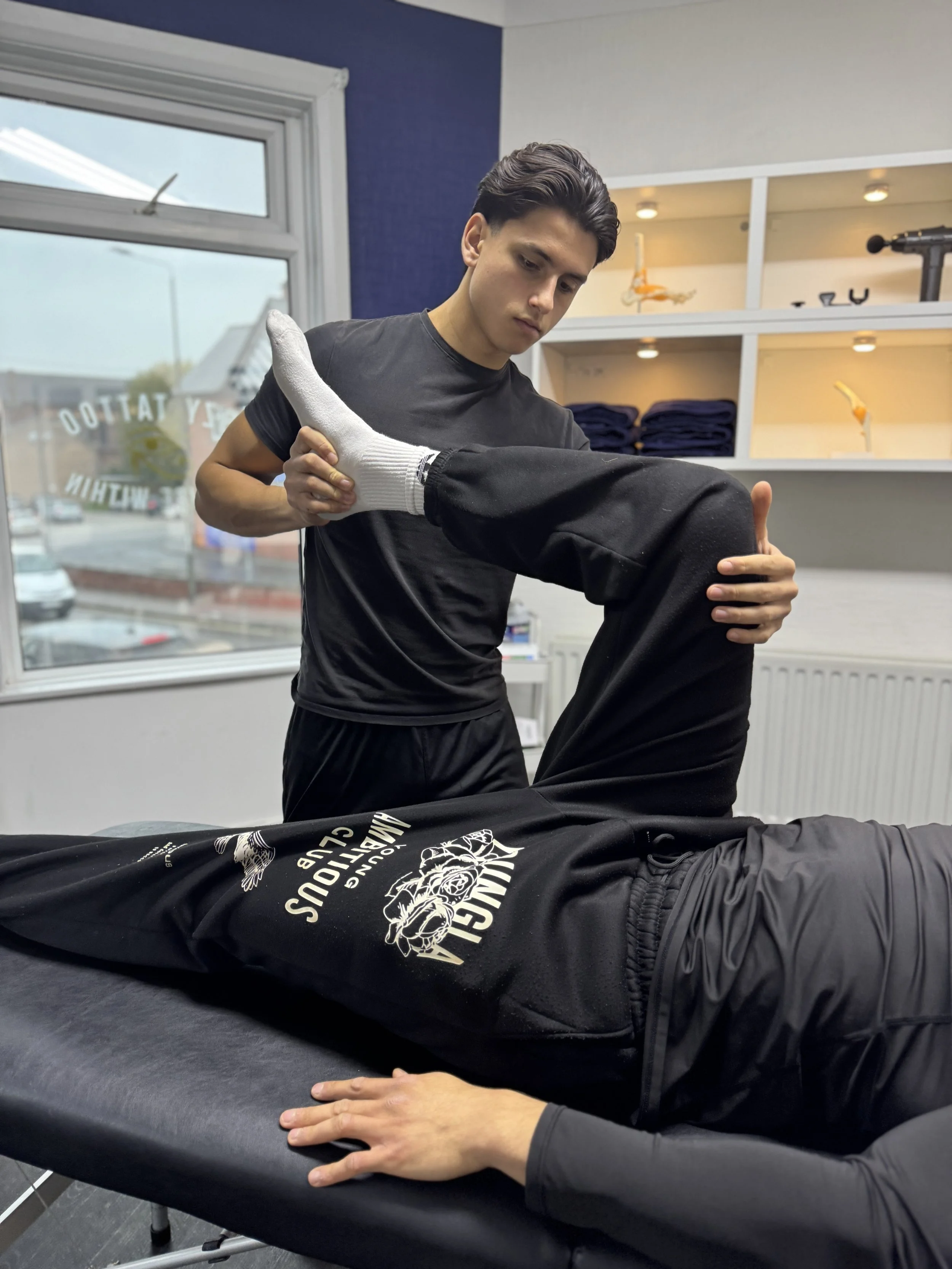 A sports therapist performing a physical assessment on a patient, holding the patient's legs while the patient is lying down.