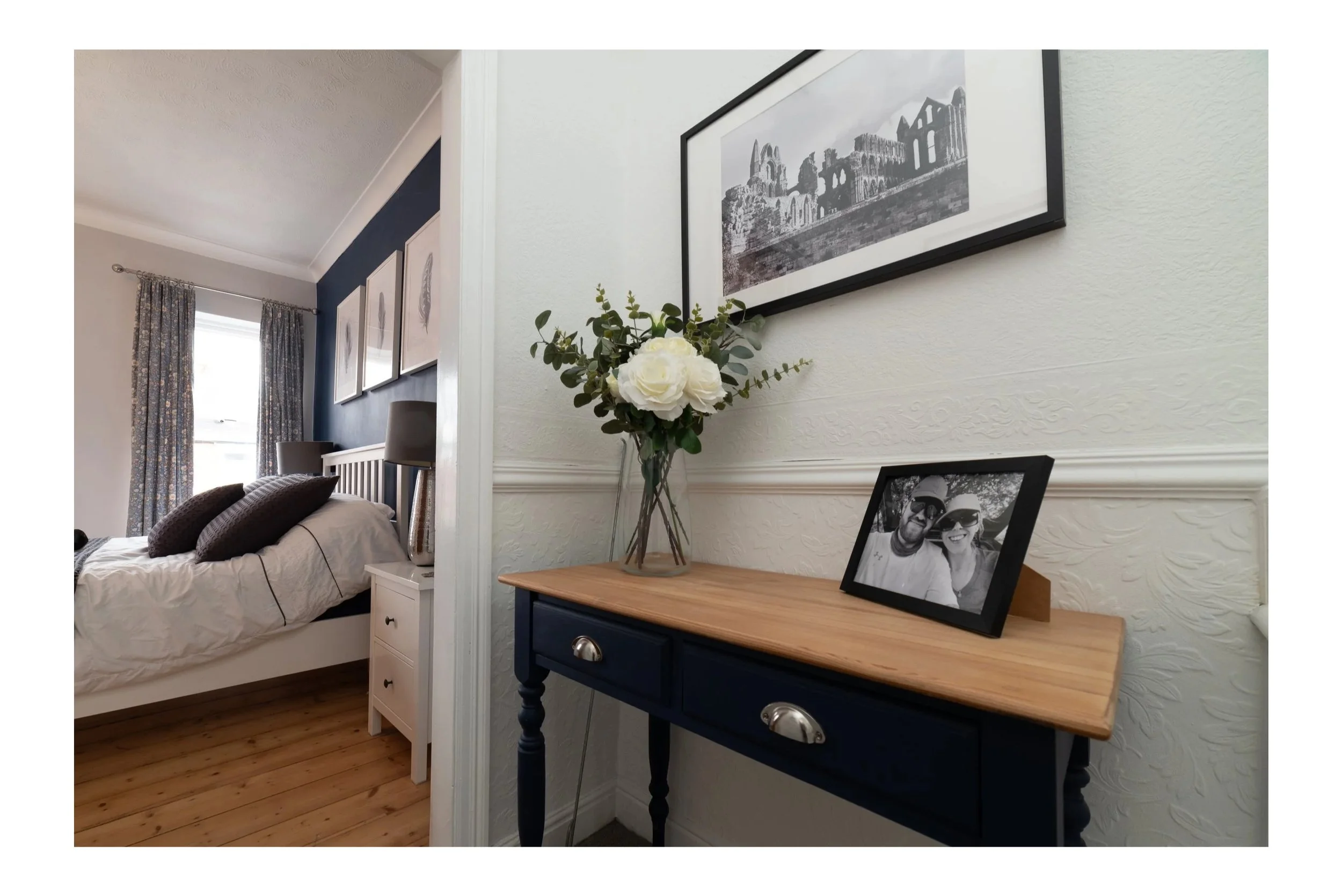 An interior view of a bedroom showing a white bed with dark pillows, a bedside table with lamps, and wall art, with a partial view of a hallway that has a side table with a flower vase and framed photograph, and artwork above it.