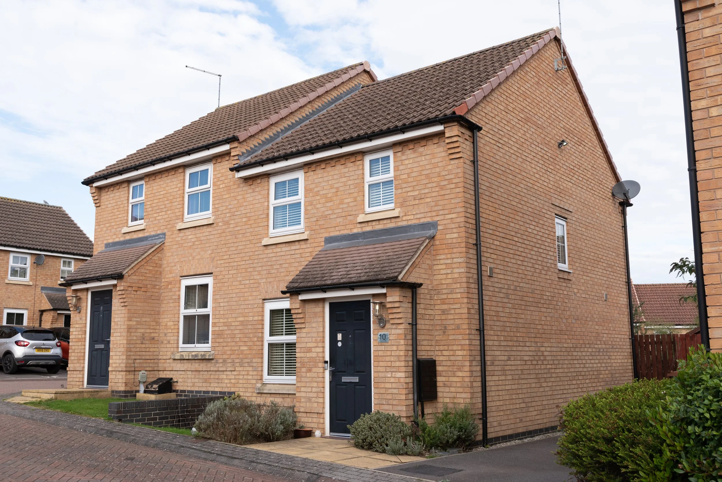 A row of modern brick townhouses with dark door entrances and small front gardens, parked cars, and a cloudy sky.