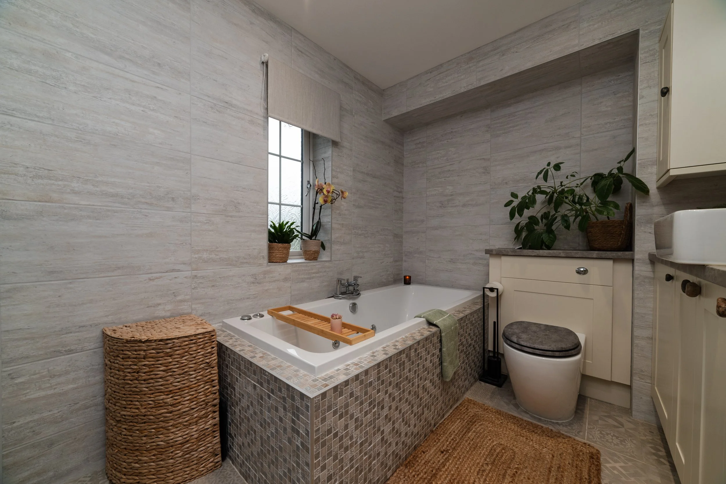 Bathroom with a small bathtub, potted plants on the window sill, wicker basket, woven mat, and a toilet