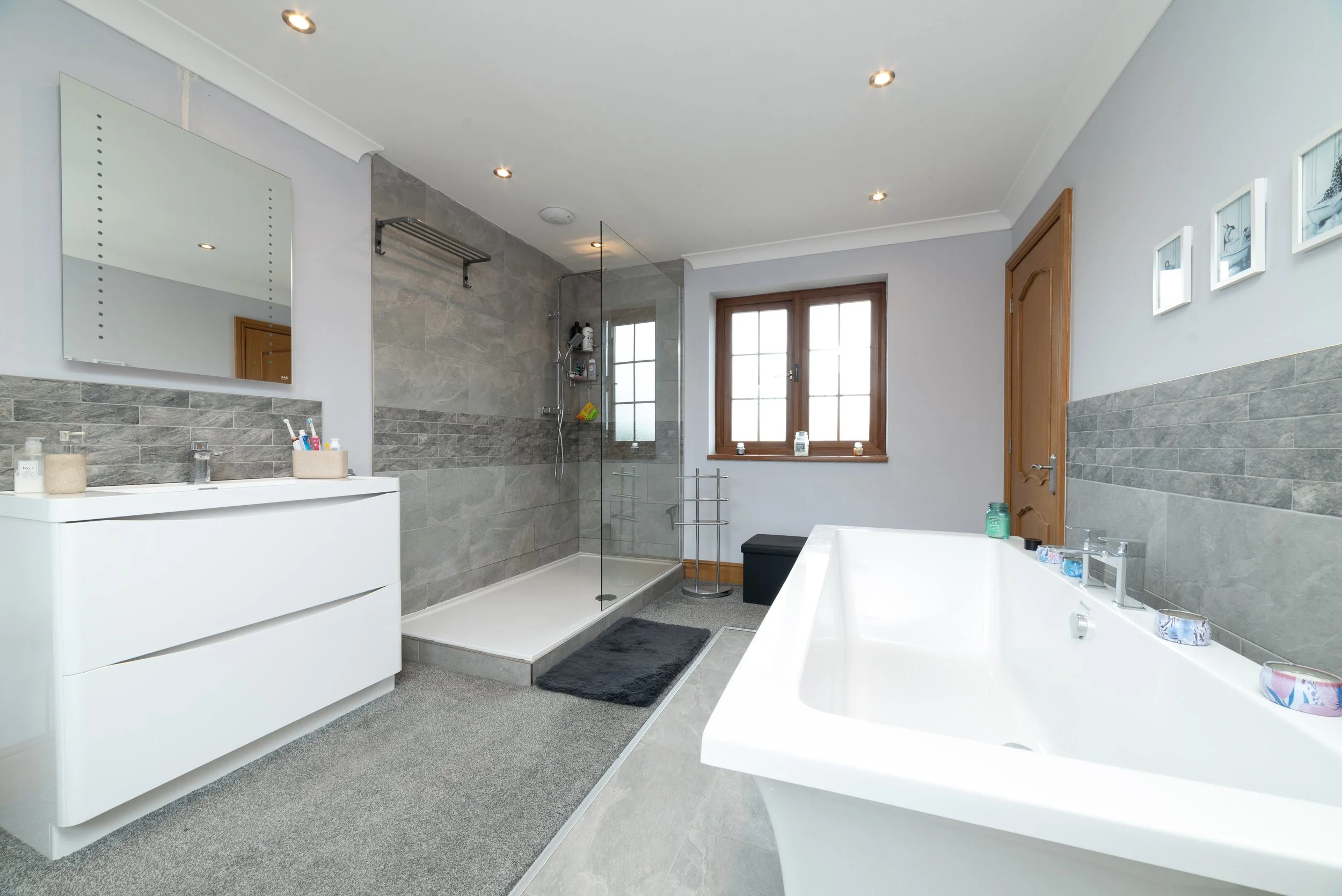A modern bathroom featuring a white bathtub, a walk-in shower with gray tiles, a black bench near a window, a white vanity with toiletries, a large wall mirror, and three small framed pictures on the wall.
