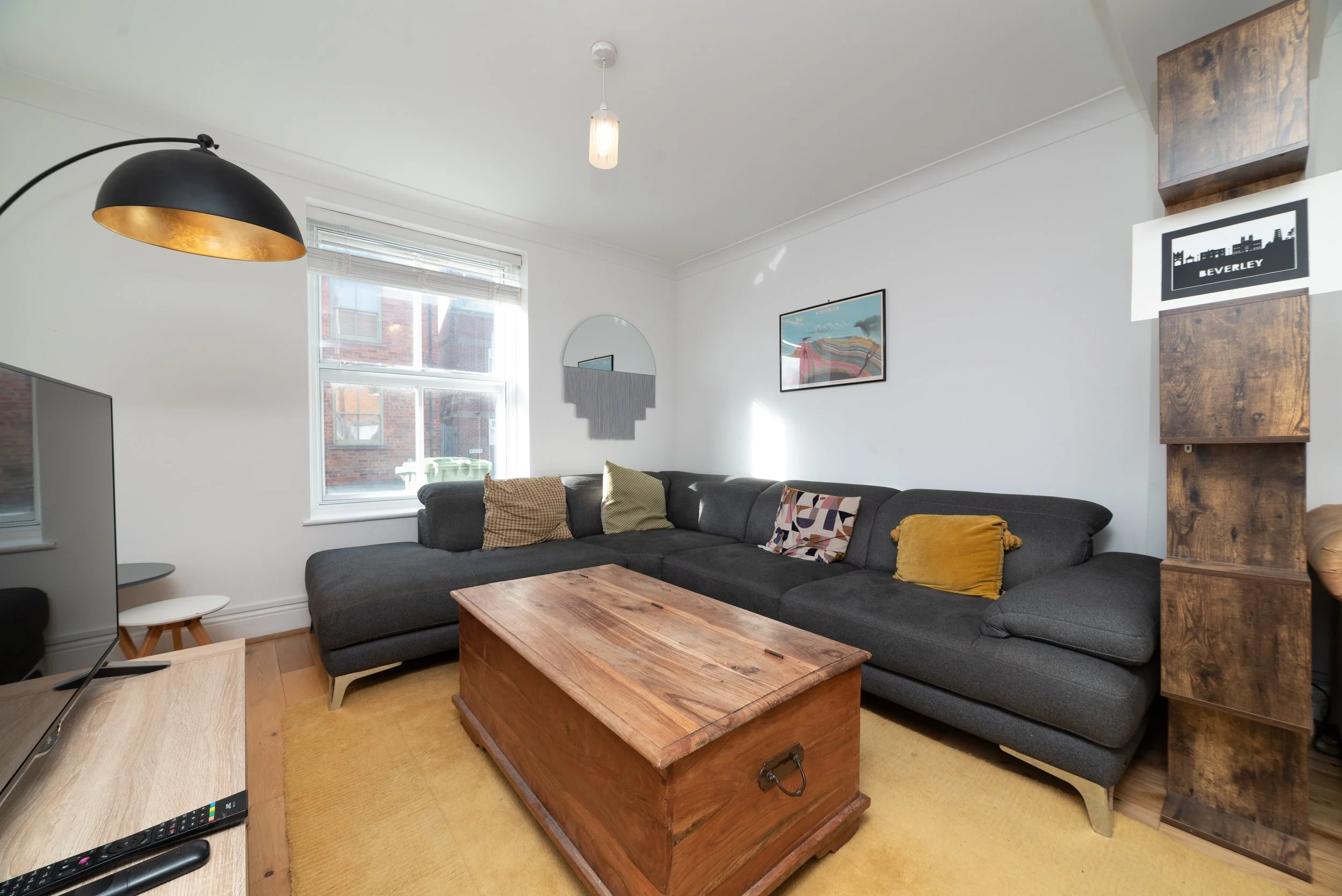 Living room with a large dark gray sectional sofa adorned with patterned and solid throw pillows, a wooden coffee table, a yellow area rug, a window with blinds, wall art, a black and gold floor lamp, a flat-screen TV, and a wooden shelving unit.