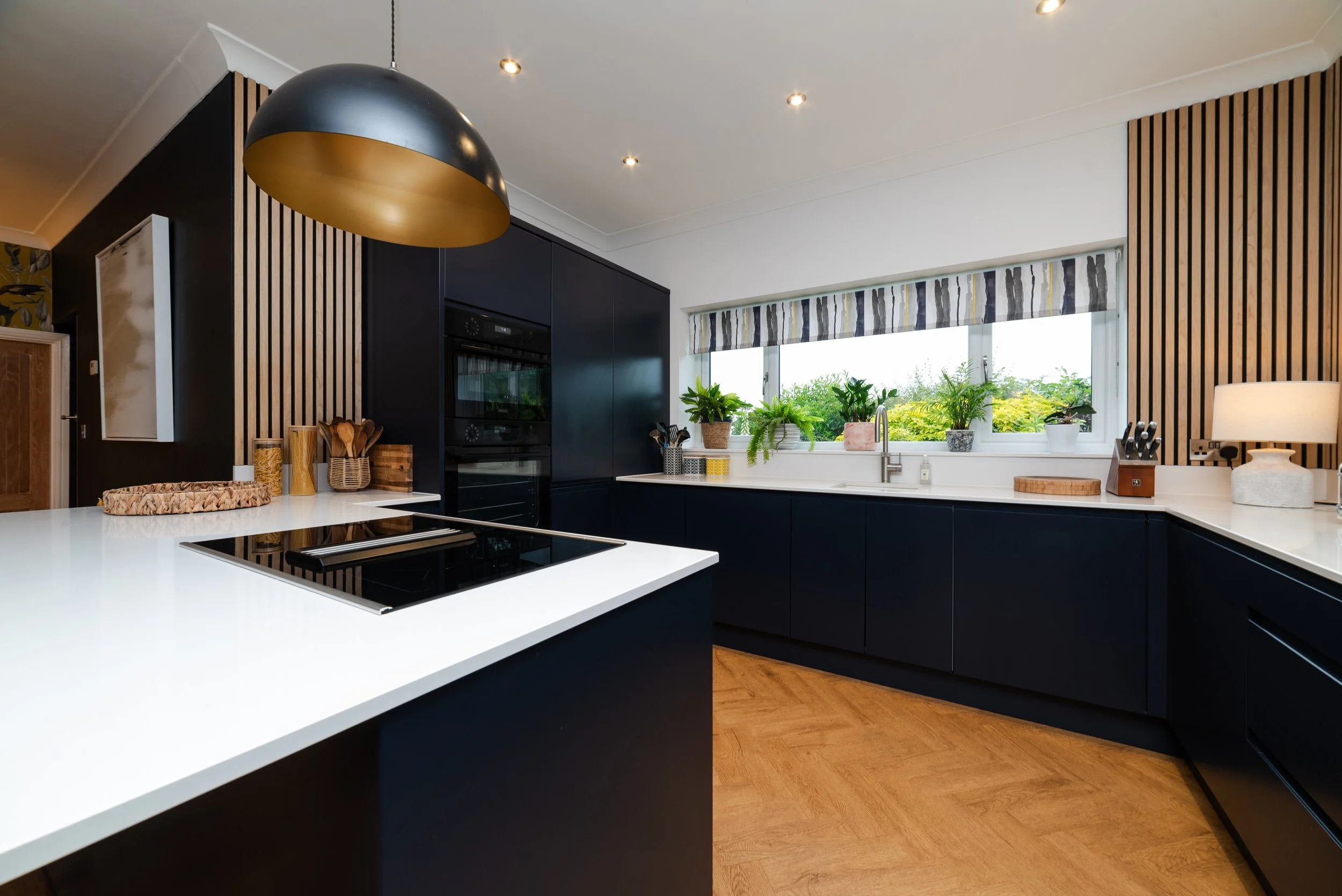 Modern kitchen with black cabinets, white countertops, a window with plants, and a black and gold pendant light.