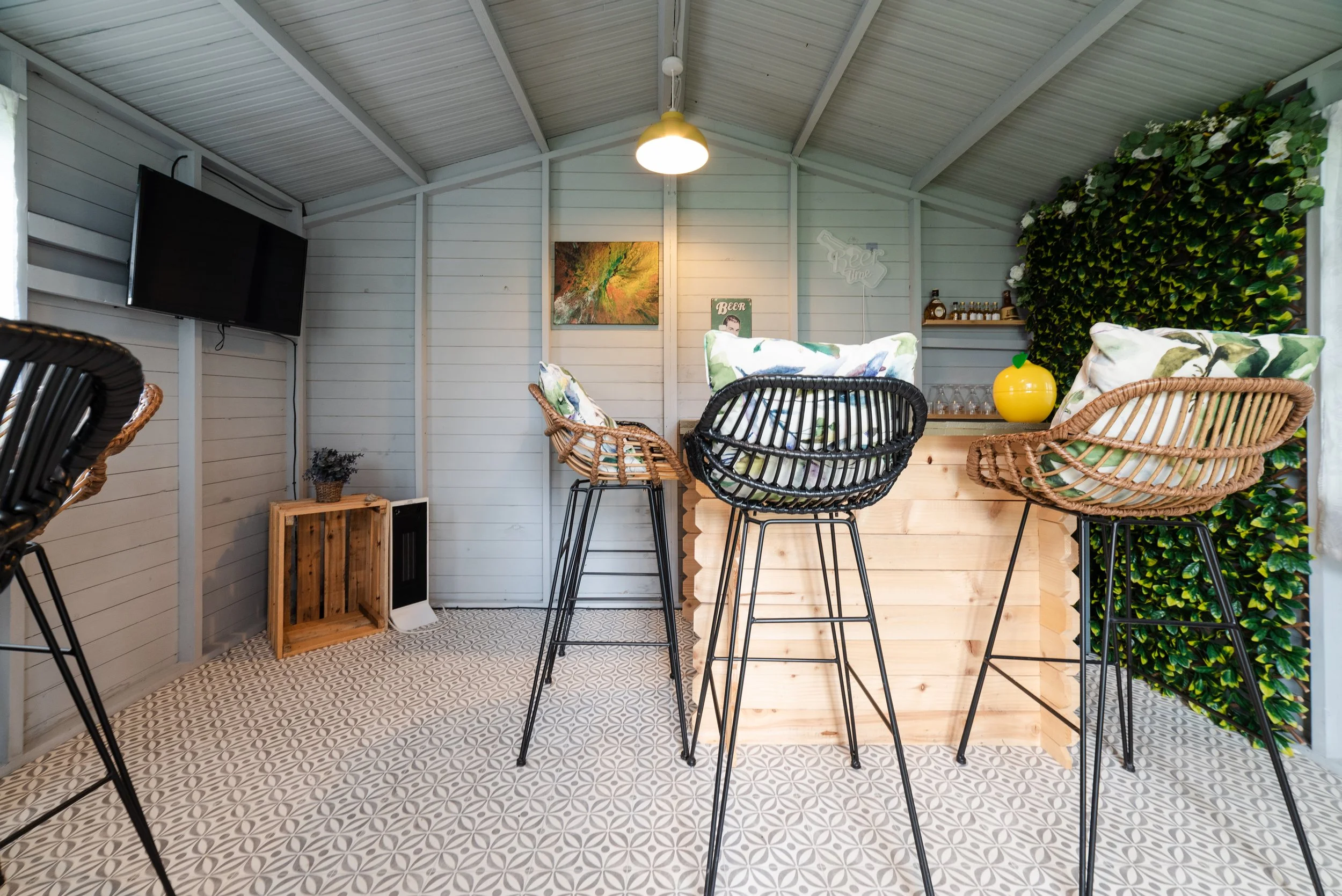Interior of a cozy bar or lounge with white paneled walls, a flat-screen TV, colorful artwork, a wooden bar counter with four barstools with leafy cushions, a large yellow apple-shaped decor, and a lush green wall on the right side.