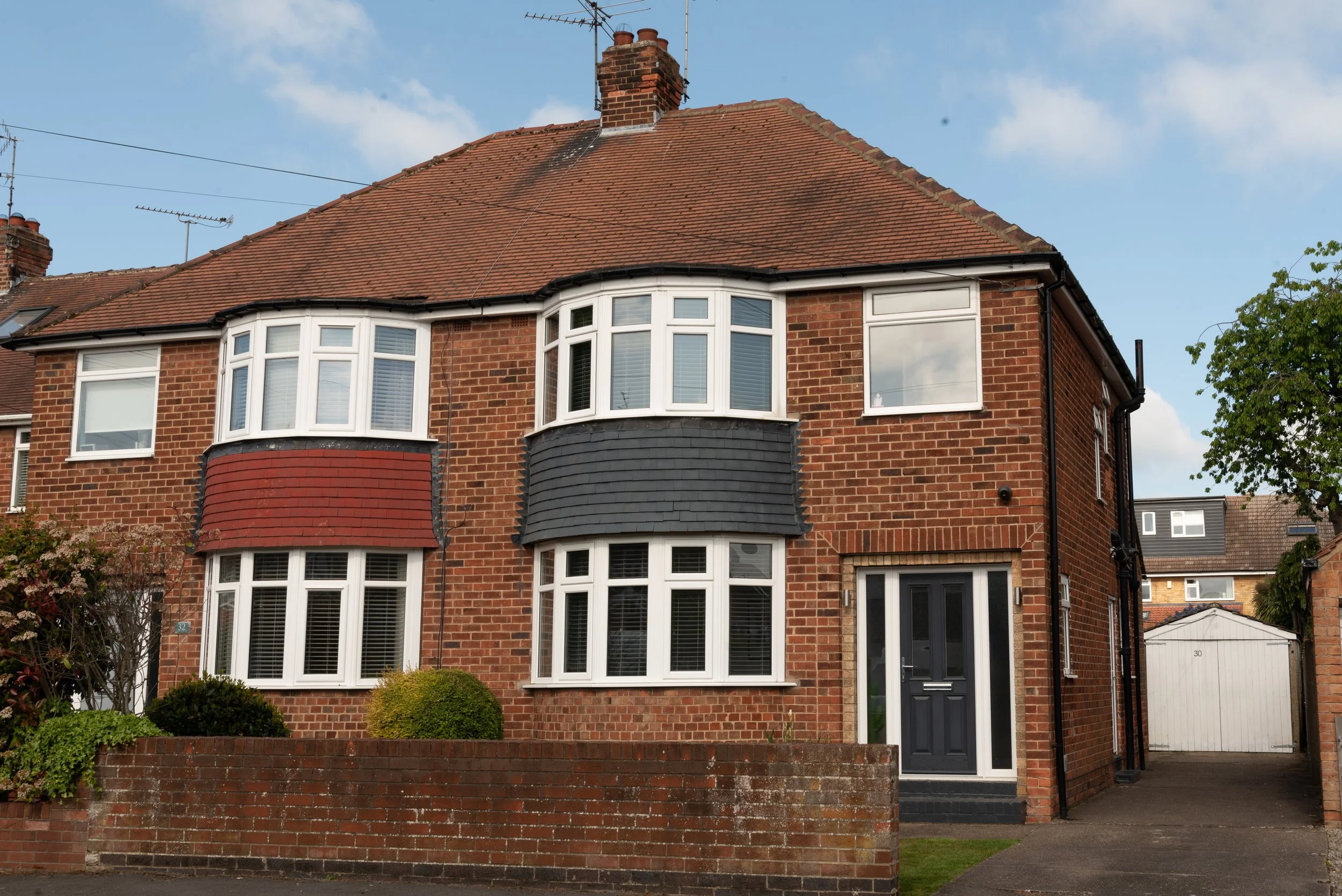 A brick house with a tiled roof, white-framed bay windows, and a dark-colored front door. There is a brick wall with bushes in front and a driveway leading to a detached garage.