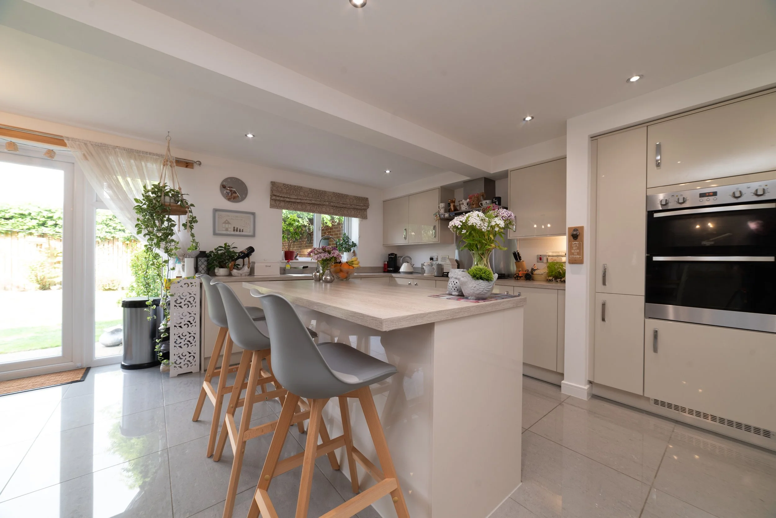 Kitchen with beige cabinets, a kitchen island with bar stools, a window with a beige valance, potted plants, flowers on the island, and stainless steel appliances, including a double oven.