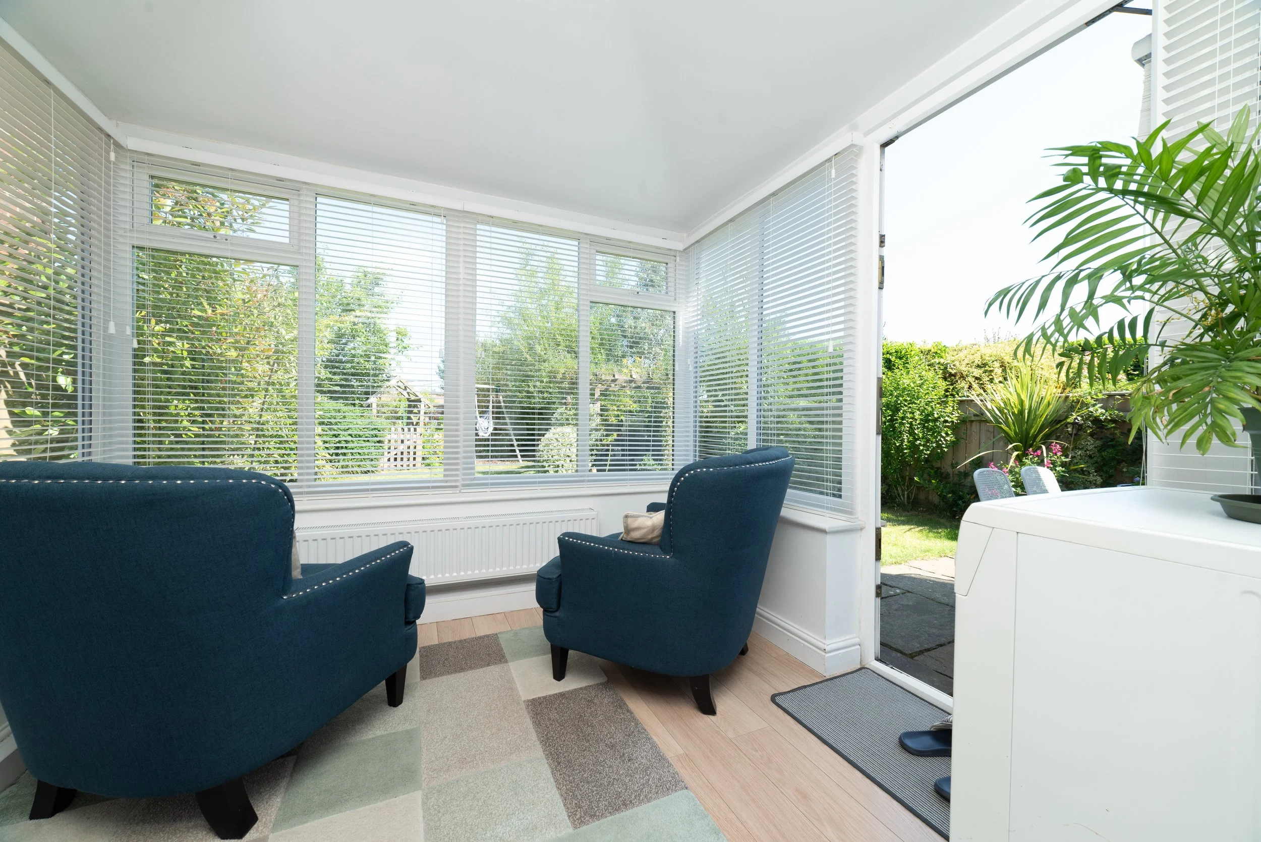 Sunlit enclosed porch with two blue armchairs, large windows with white blinds, and a door leading outside to a garden with plants and outdoor furniture.