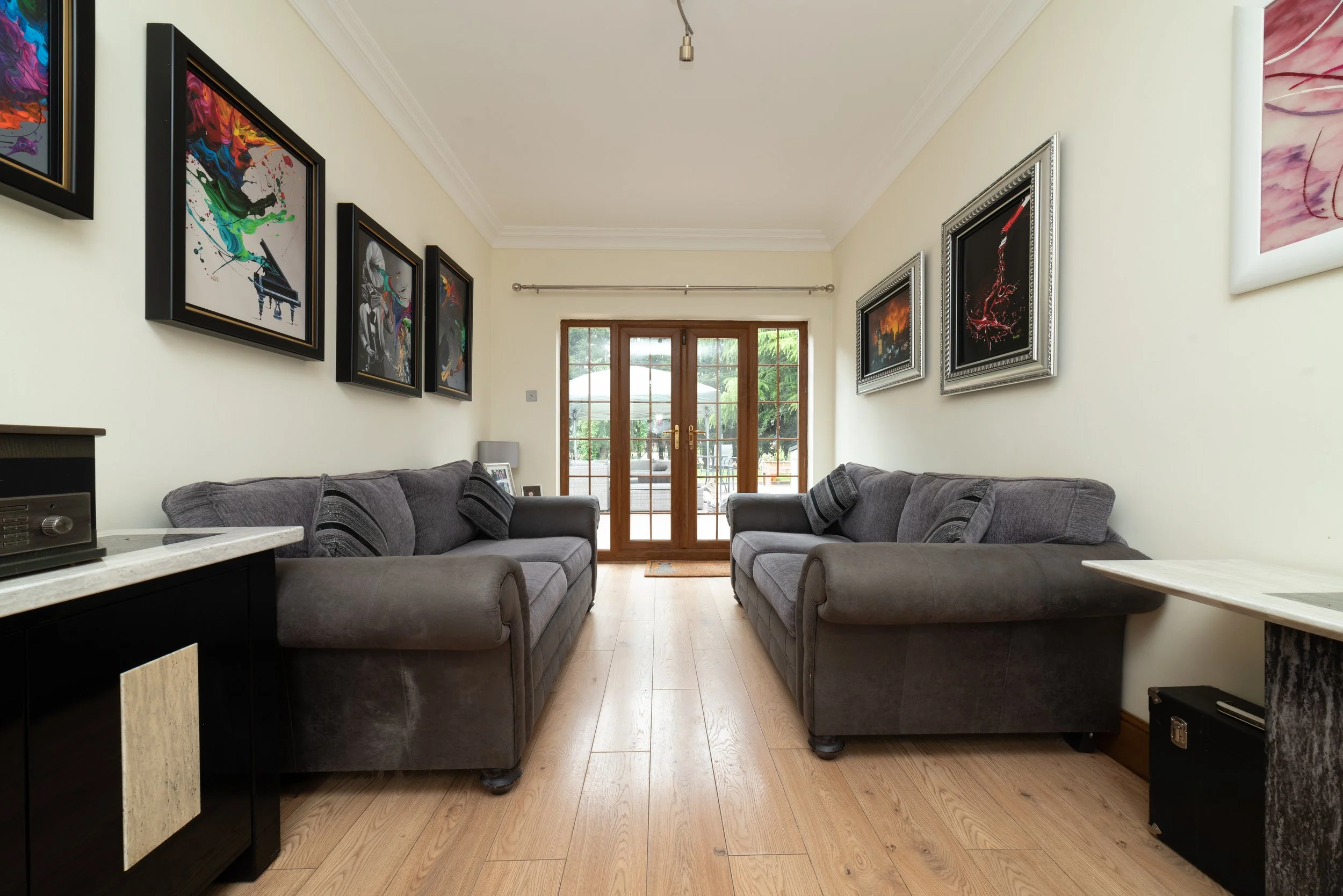 Living room with two grey sofas facing each other, wooden floor, glass double doors leading outside, and framed artwork on the walls.