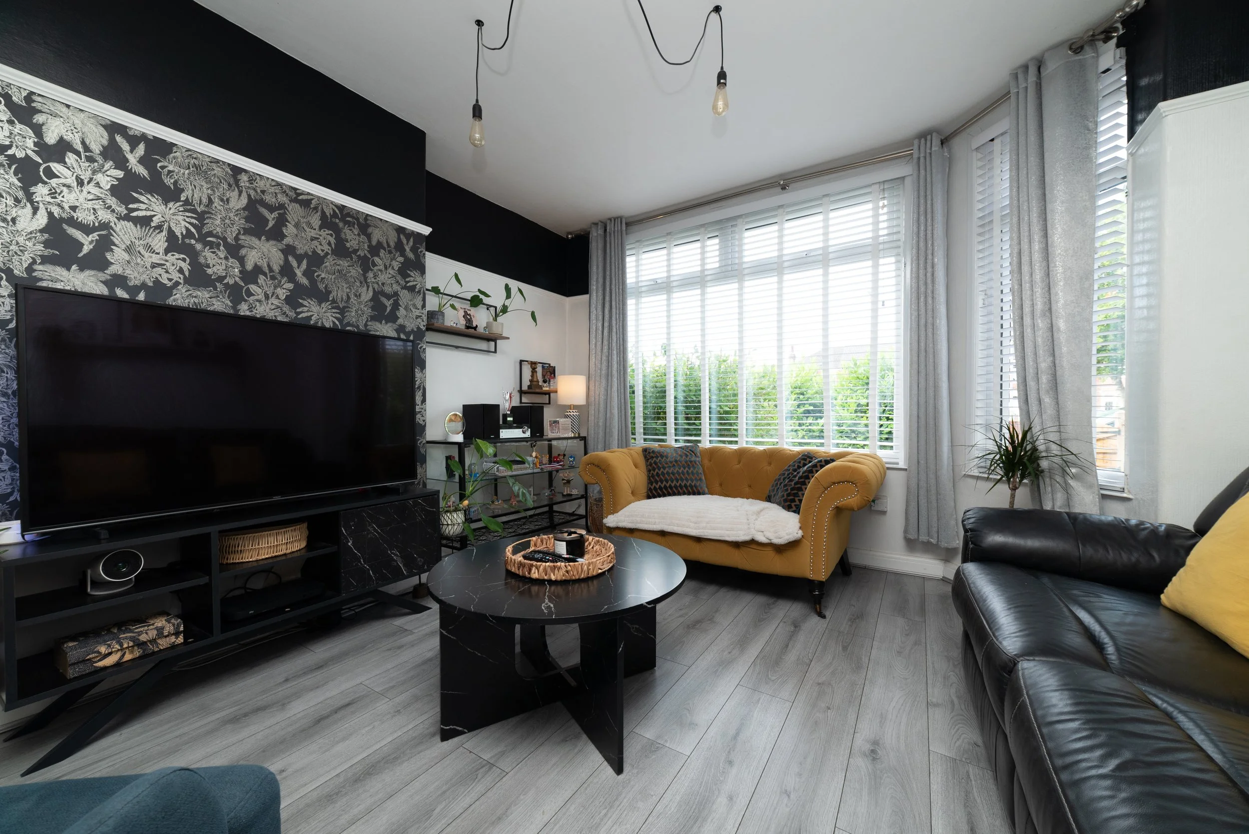 Living room with large window, yellow tufted sofa, black leather sofa, round black marble coffee table, TV on black stand, patterned wallpaper behind TV, gray flooring, and potted plants.