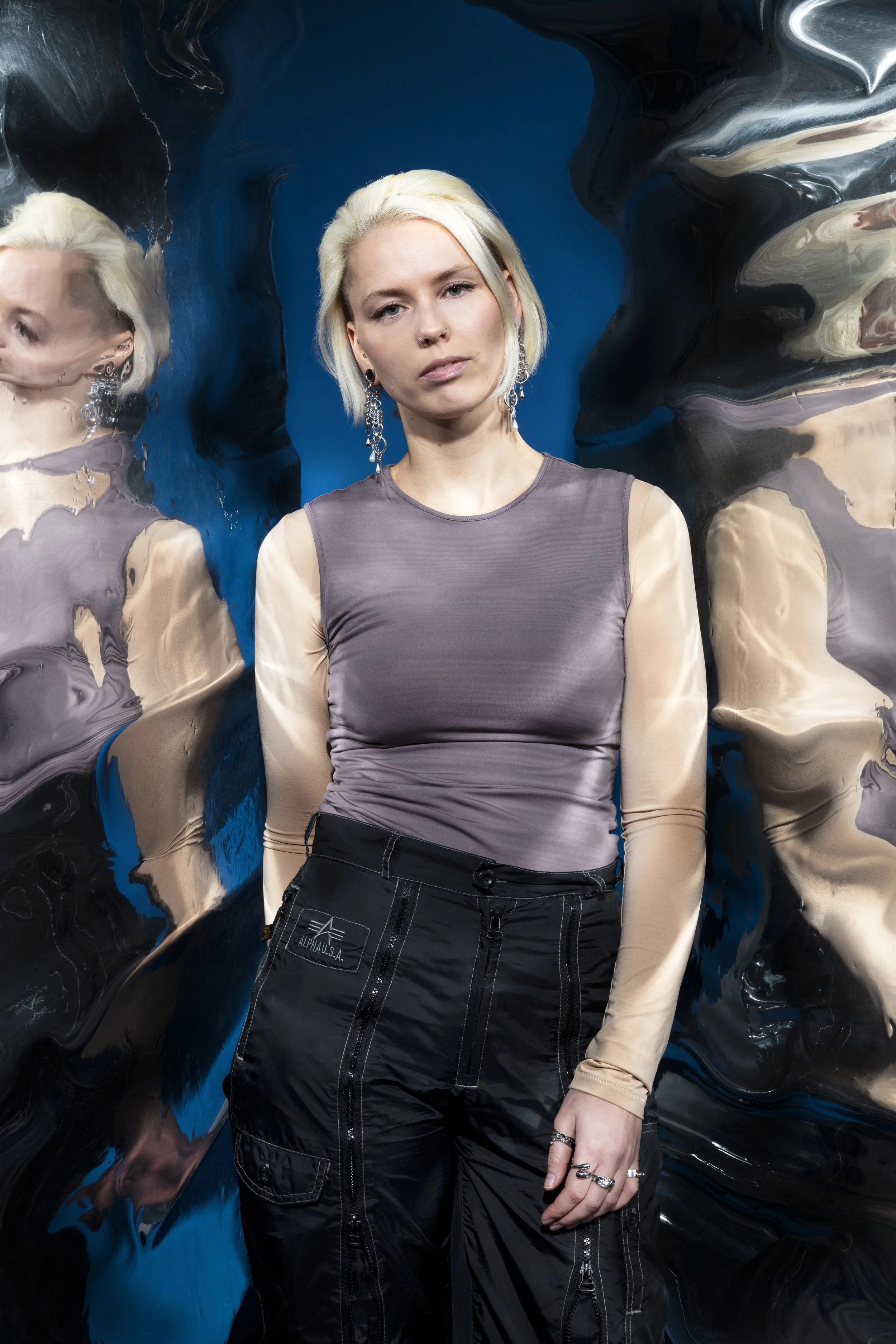 Een vrouw met blond haar en sieraden poseert voor een watervervormende spiegel die haar weerspiegelen vervormt.