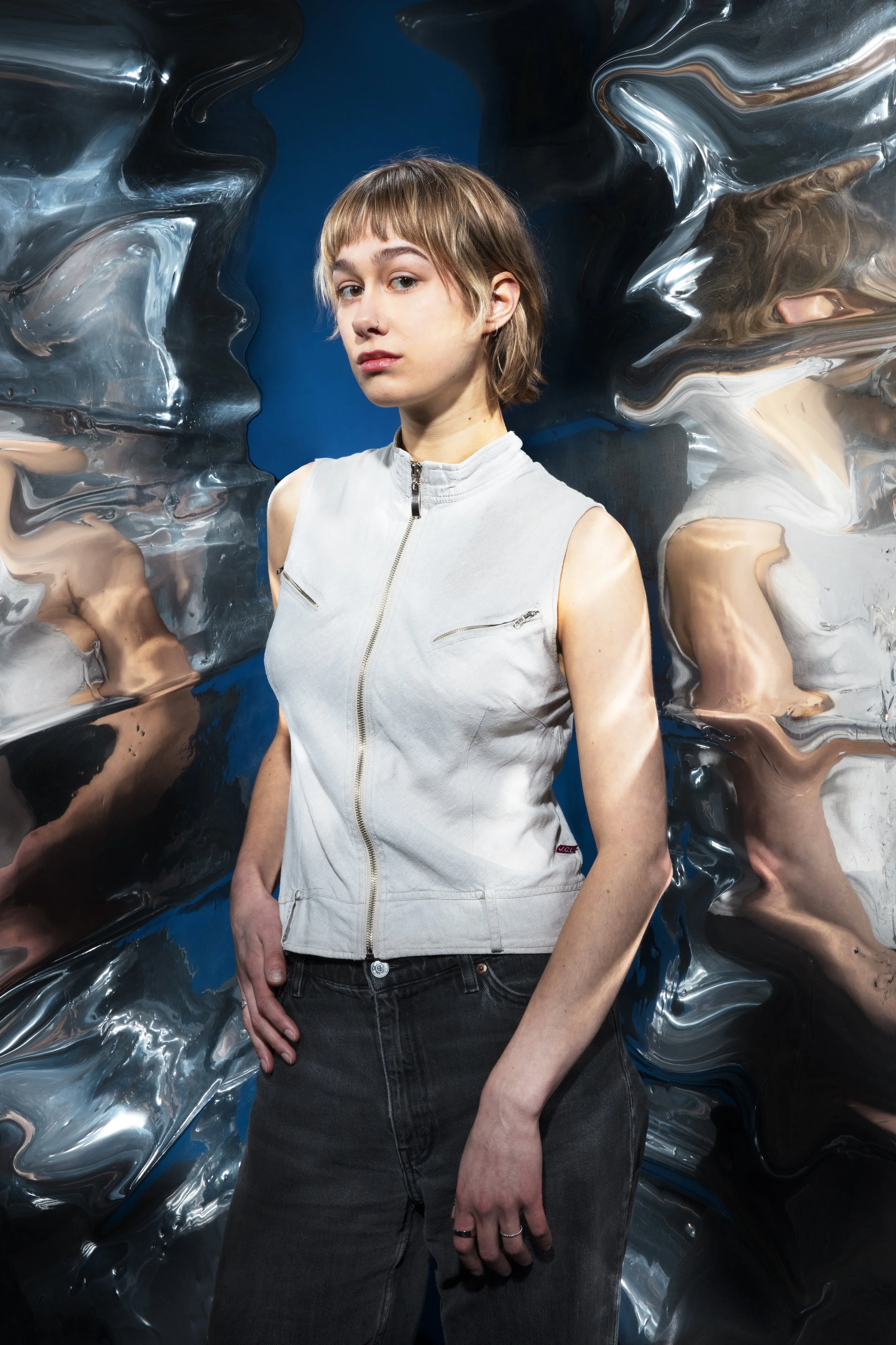 Een jonge vrouw met kort bruin haar staat voor een kunstzinnige, reflecterende achtergrond met golvende metallic patronen.