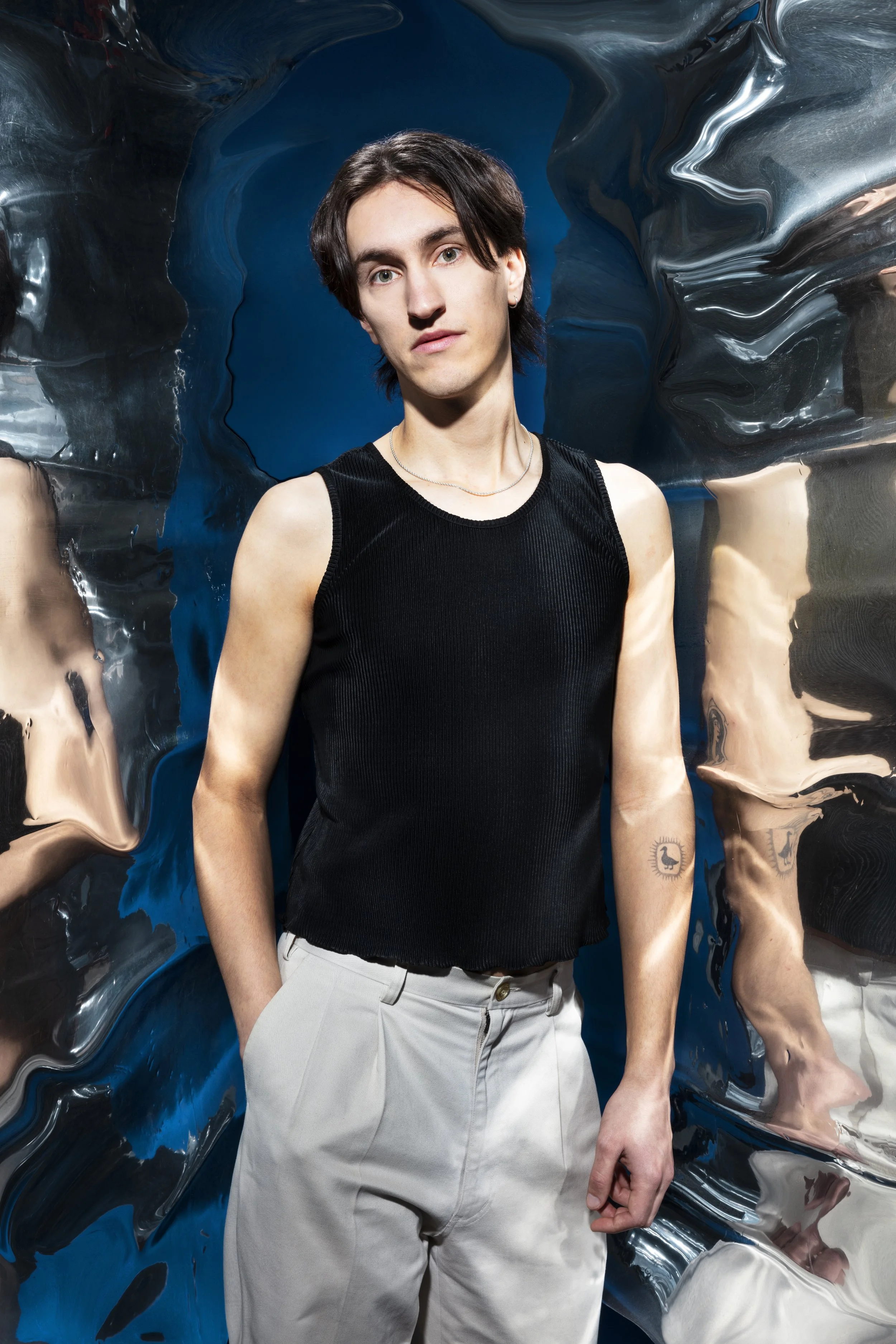 Portret van een jonge man met zwart haar, licht getint gezicht, in een zwart mouwloos shirt en witte broek, staand voor een gestructureerde, reflecterende achtergrond.