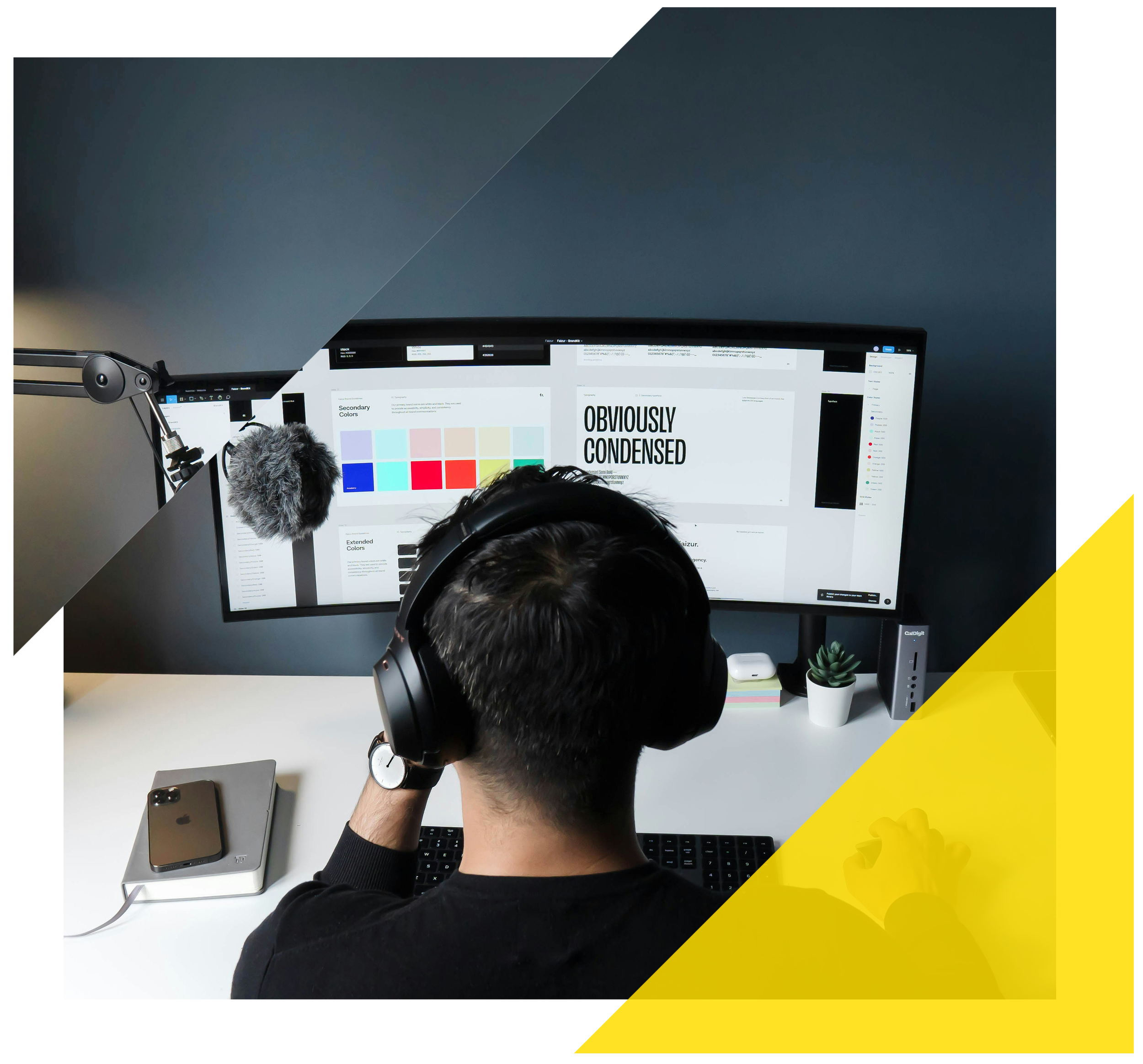 Person wearing headphones working at a computer with dual monitors, on white desk with smartphone, tablet, potted plant, and office accessories, in a dark-colored room.