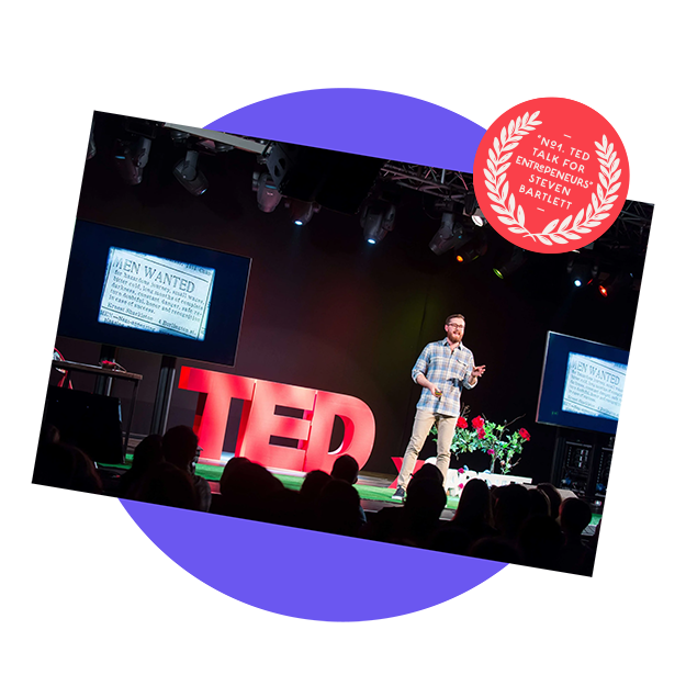 A man giving a TED Talk on stage with large TED letters behind him, presentation slides on screens, and an audience watching.