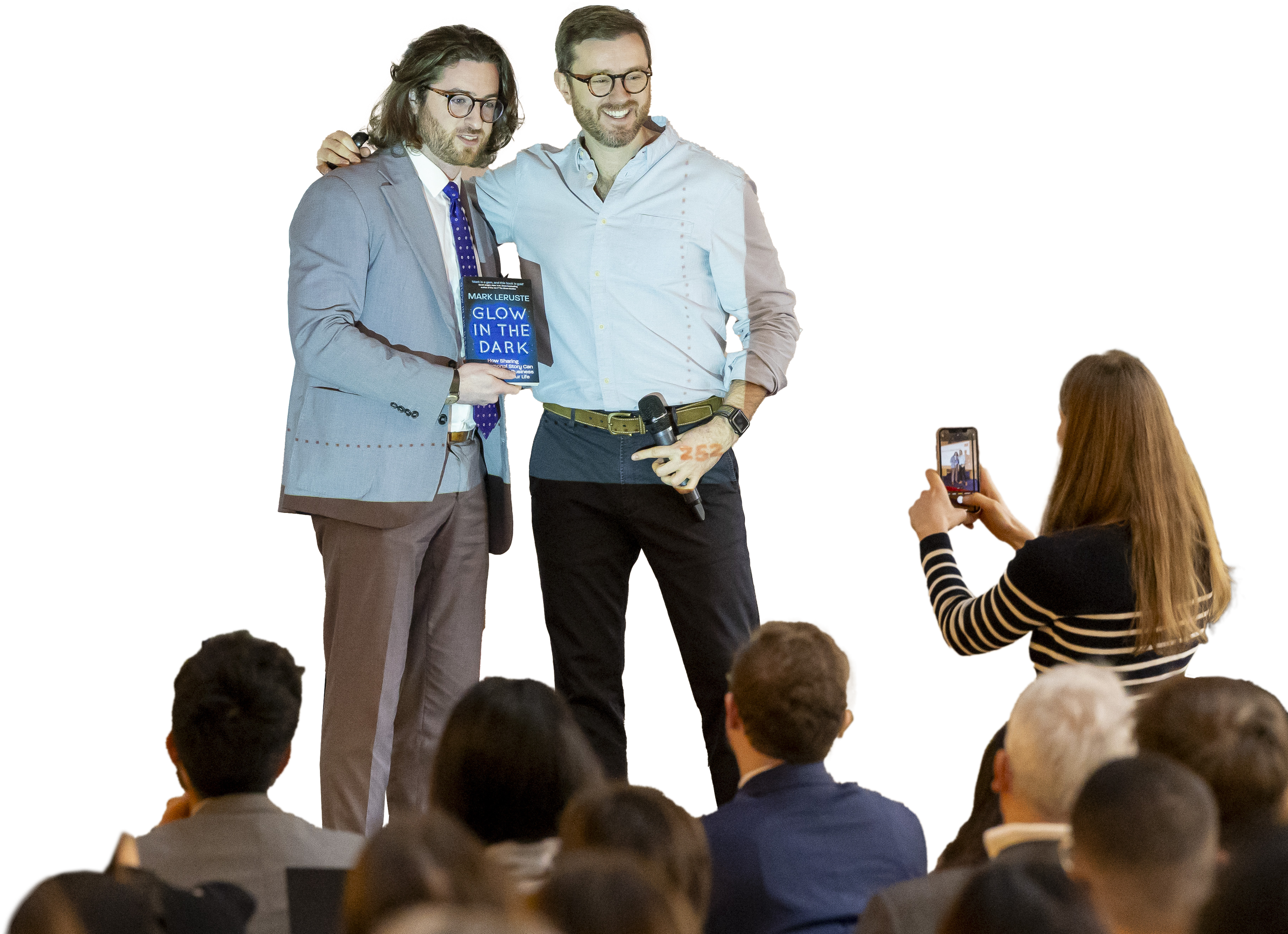 Two men standing on stage, one holding a book titled 'Glow in the Dark,' and the other holding a microphone, while a woman takes a photo with her phone in front of an audience.