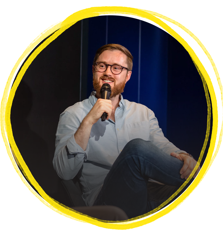 A man with glasses and a beard smiling while holding a microphone, sitting on a chair with a dark blue background.
