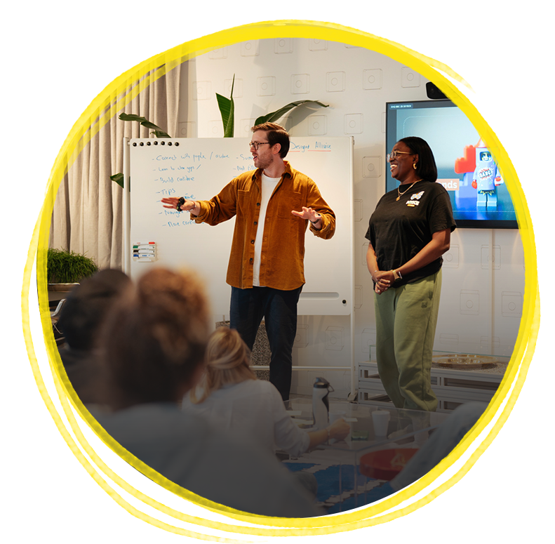 Two presenters, a man and a woman, speaking to a group during a workshop or presentation in a modern, well-lit room, with a whiteboard and a large screen behind them.
