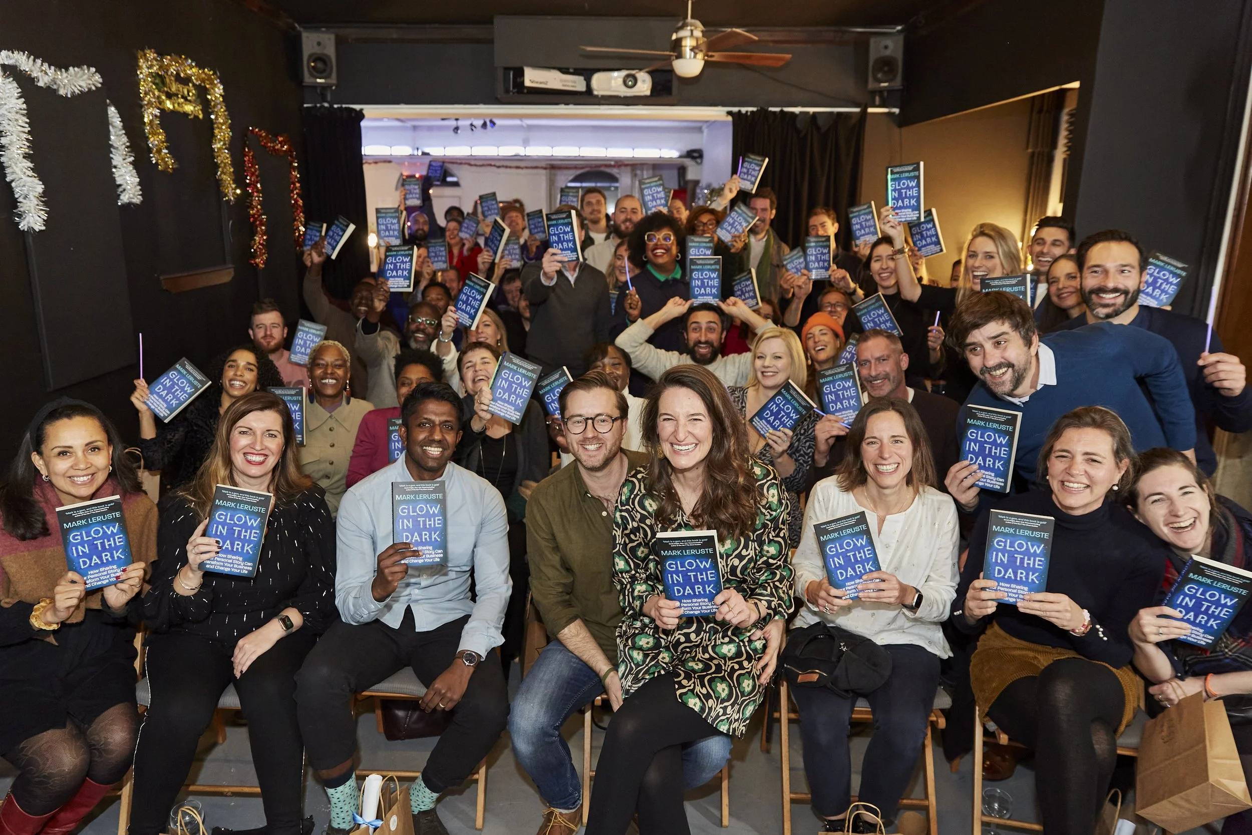 Group of people gathered in a room, holding up a book titled 'Glow in the Dark' by Mark Leruste, smiling and celebrating.