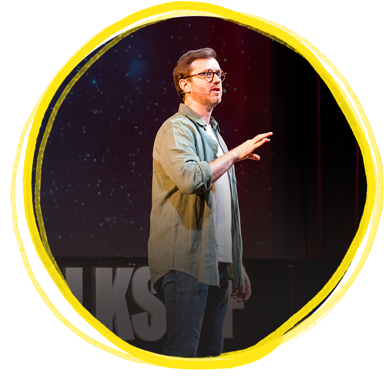 A man with glasses, wearing a light gray jacket and a white t-shirt, is speaking on stage with a dark background featuring a starry sky. He appears to be explaining something during a presentation or talk.